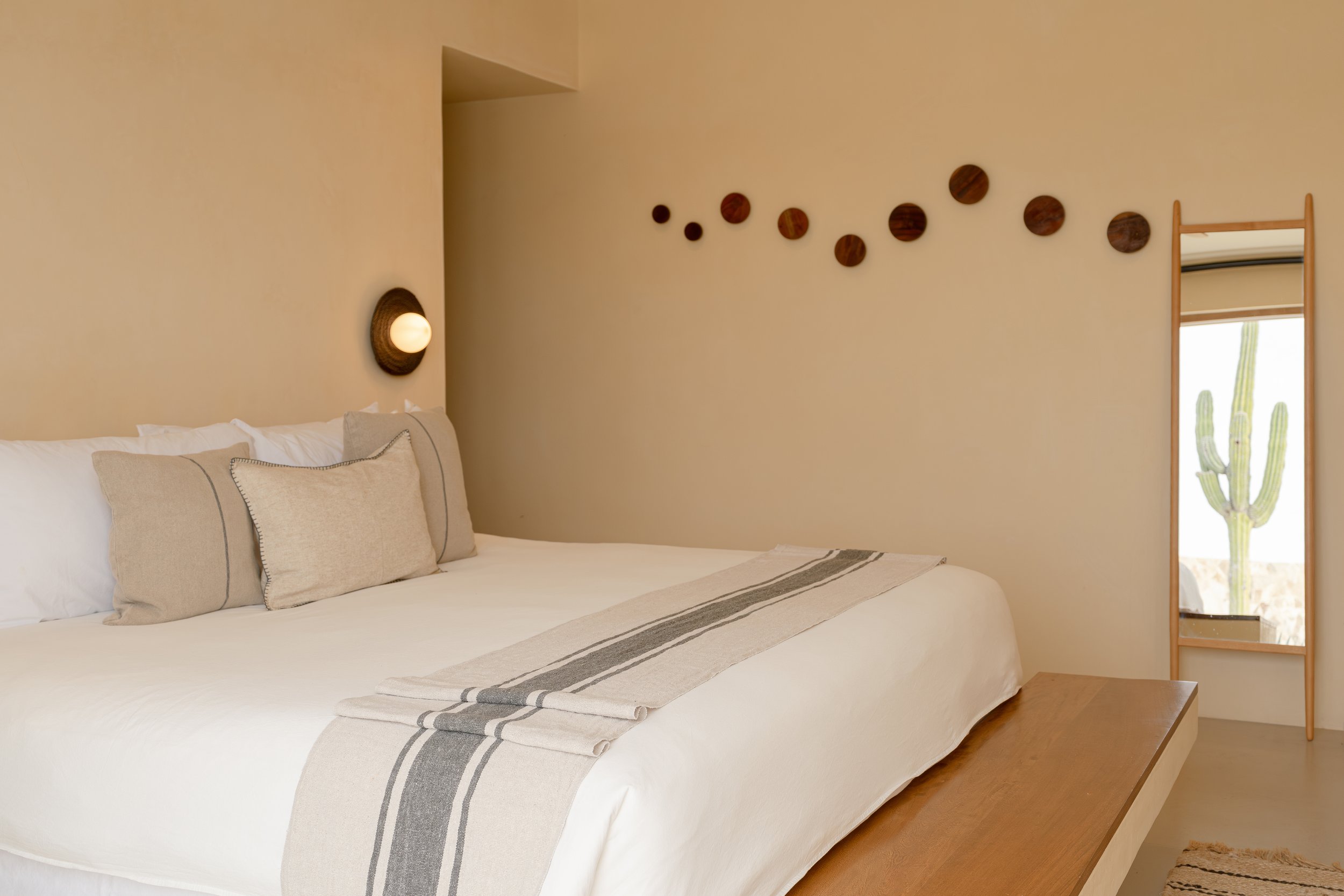 Minimalist bedroom with a large bed, beige pillows, and a striped throw. A round wall light and circular decorations adorn the wall. A wooden ladder-style mirror reflects a cactus outside.