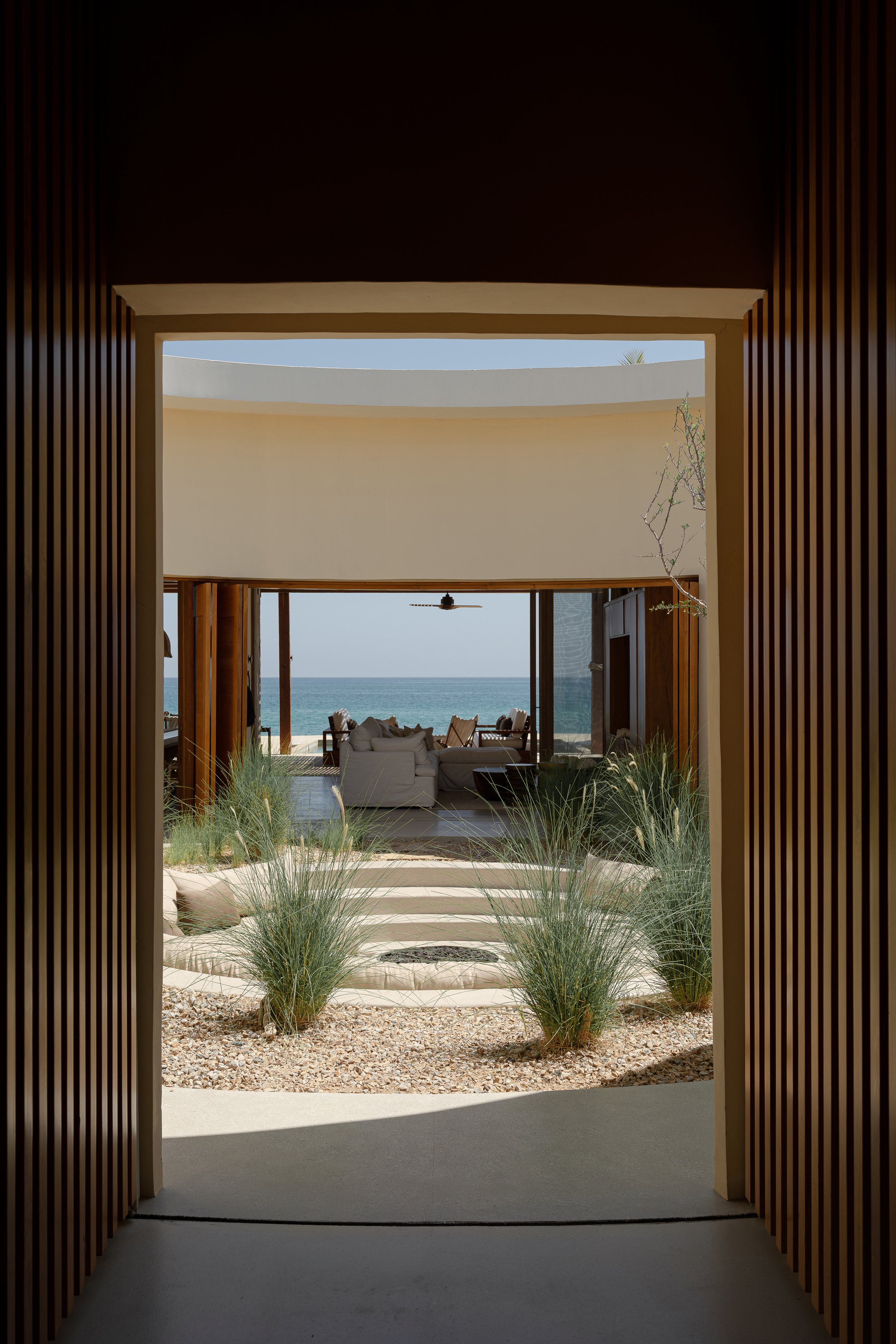 View through a corridor opening to a bright outdoor space with stone steps, ornamental grasses, and patio seating overlooking the ocean.