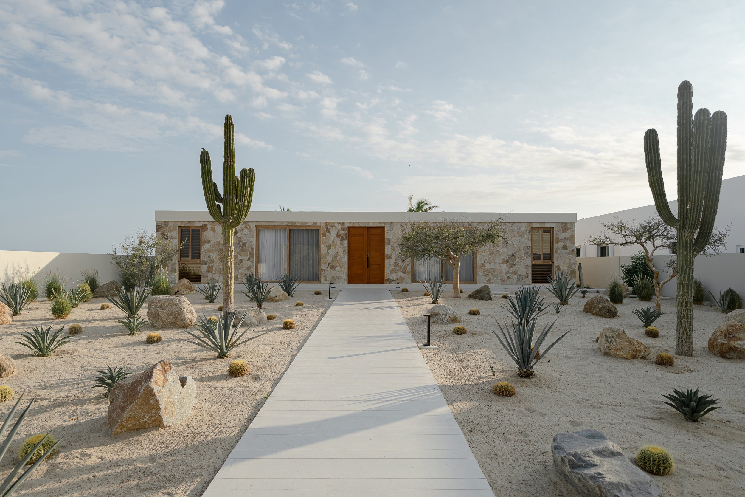 Modern desert house with stone exterior, surrounded by sand, cacti, and desert plants, featuring a central walkway leading to the entrance.