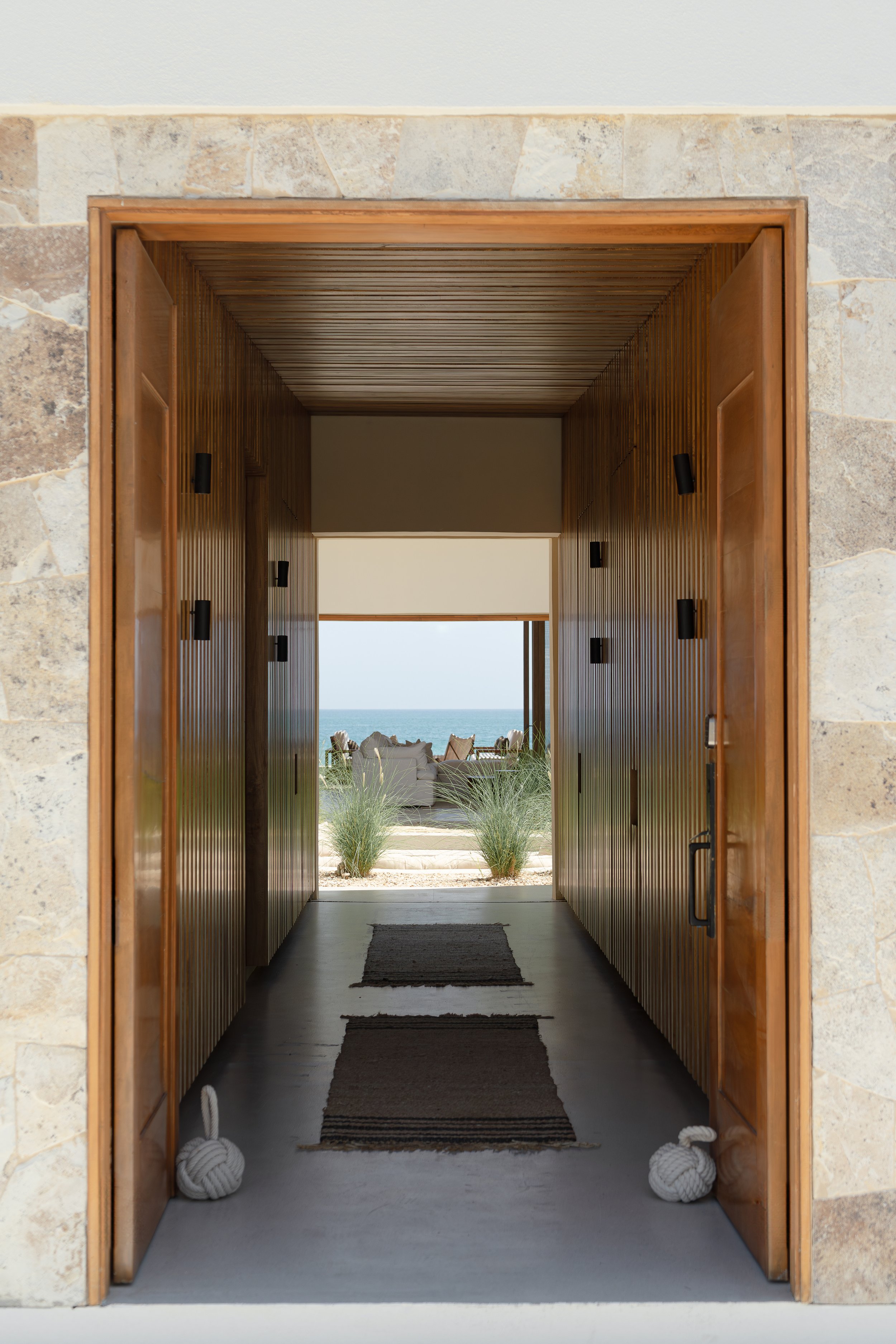 View through a modern hallway with wooden walls and ceiling, leading to an outdoor patio with a seating area and ocean view.