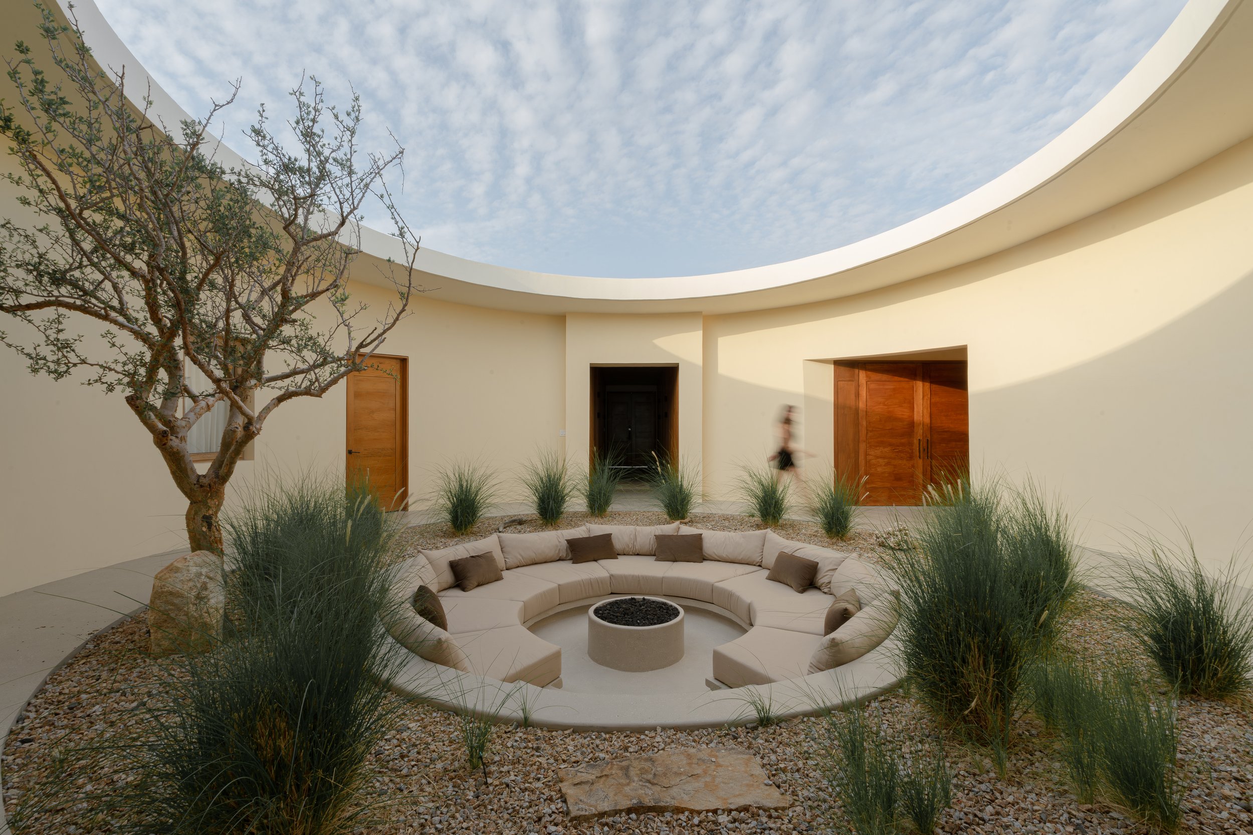 Outdoor courtyard with circular seating around a fire pit, surrounded by gravel and grass, under a partially open sky.