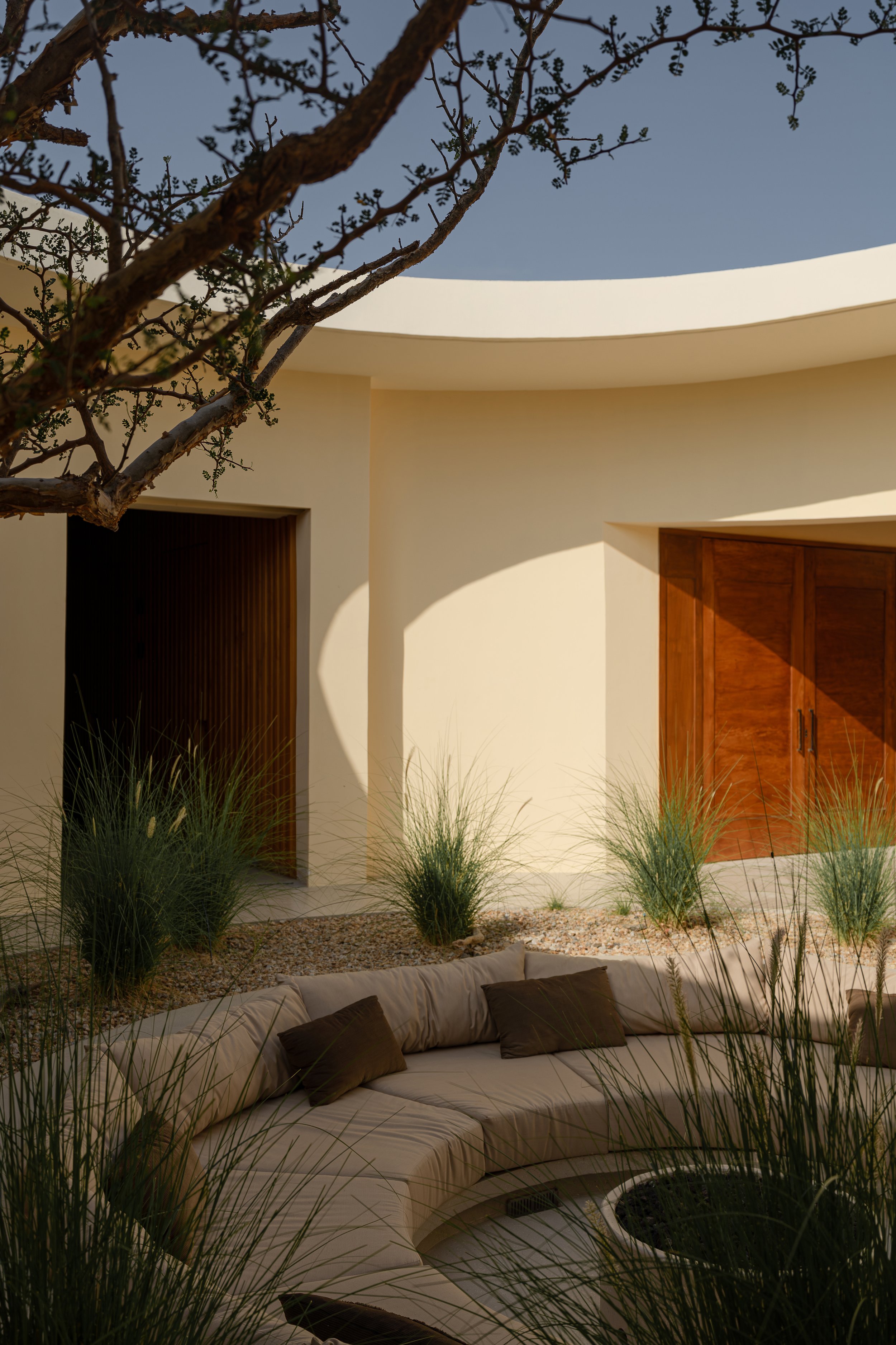 Modern outdoor patio with beige curved sofa, beige cushions, tall grasses, gravel ground, wooden double doors, and a tree branch under clear sky.