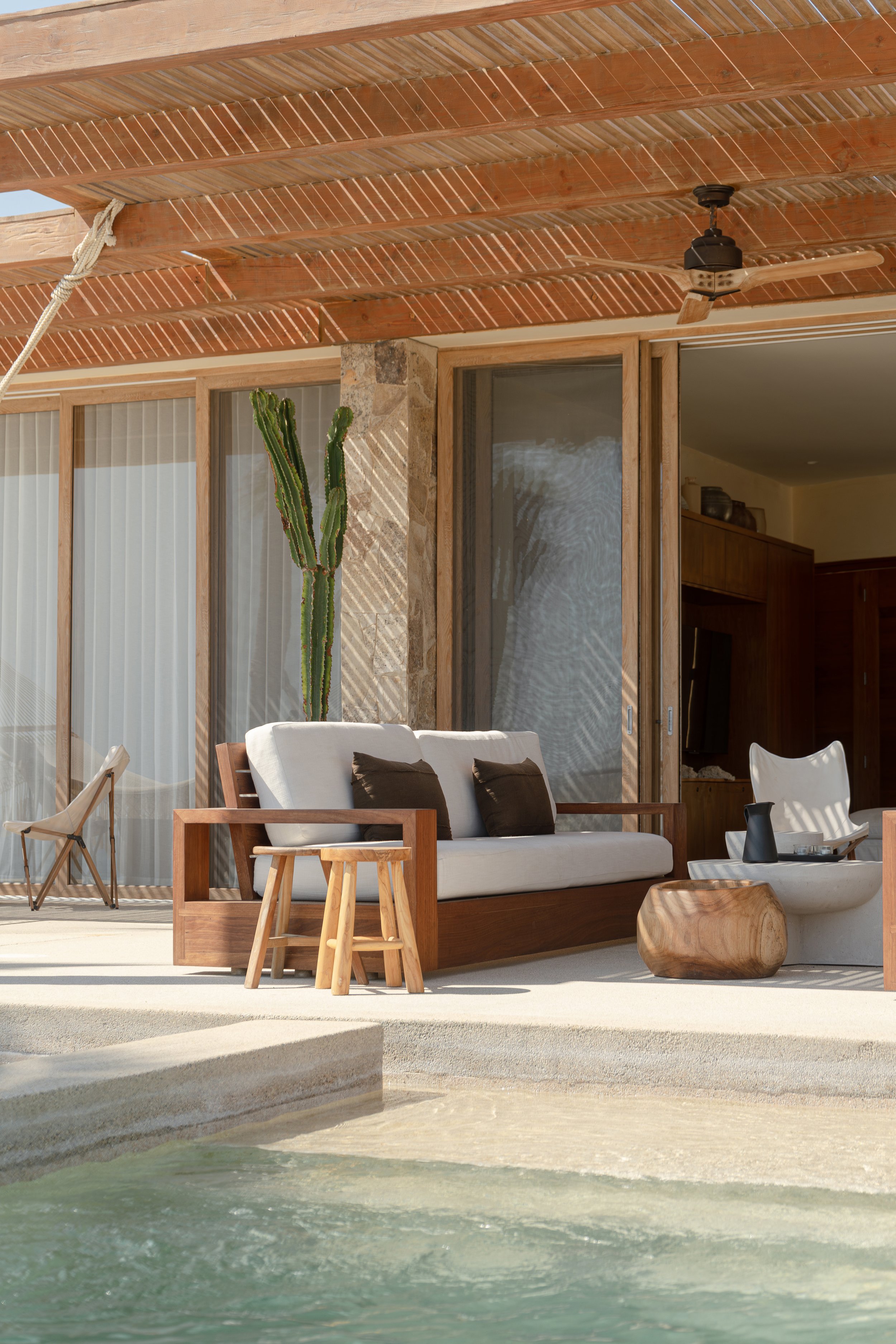 Modern outdoor patio with wooden furniture, including a sofa and table, under a pergola. Stone walls and sliding glass doors lead to an indoor area. A pool is in the foreground.