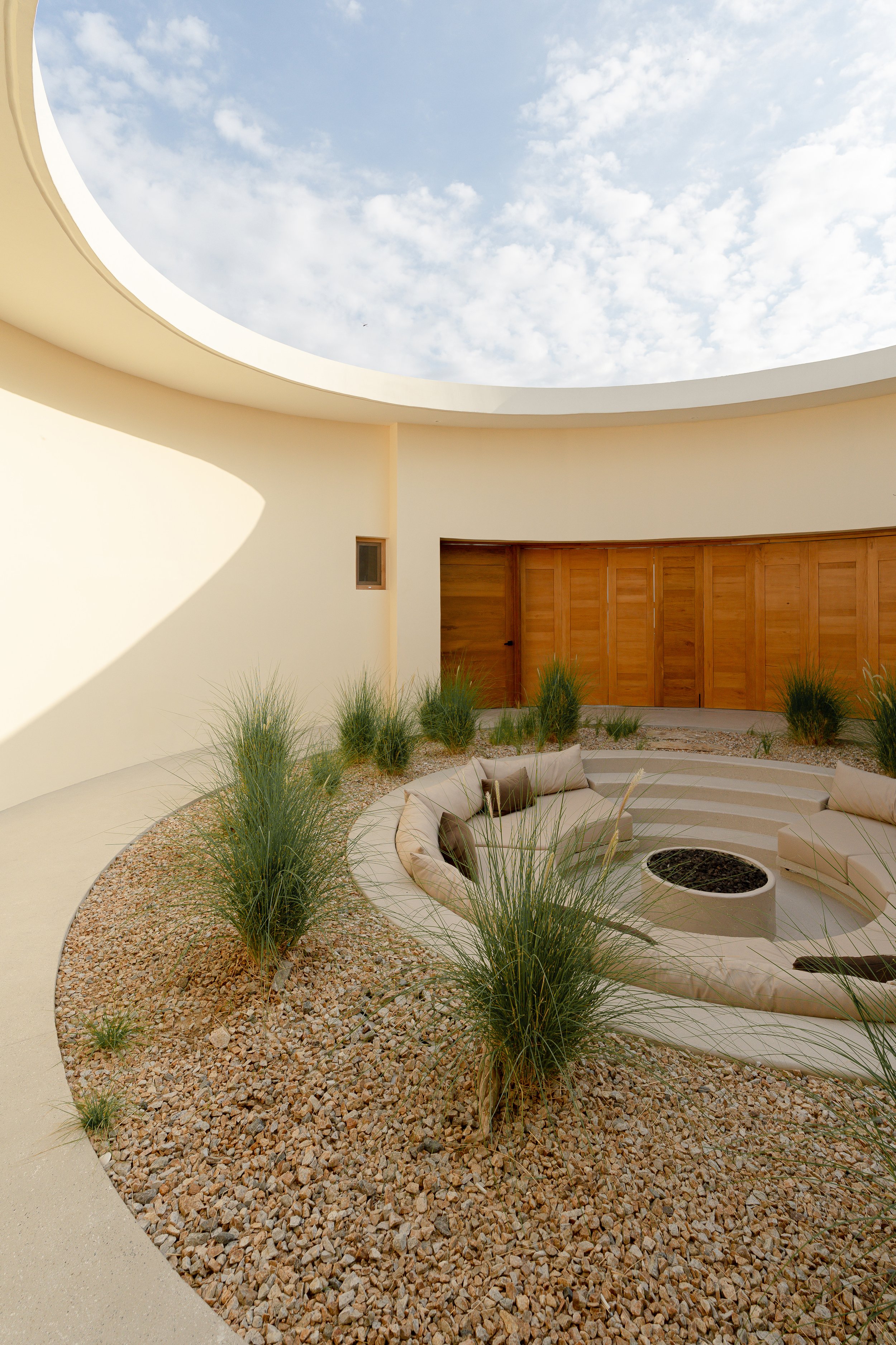 Circular open-air courtyard with gravel, grass plants, wooden doors, and a central sunken seating area with a fire pit.