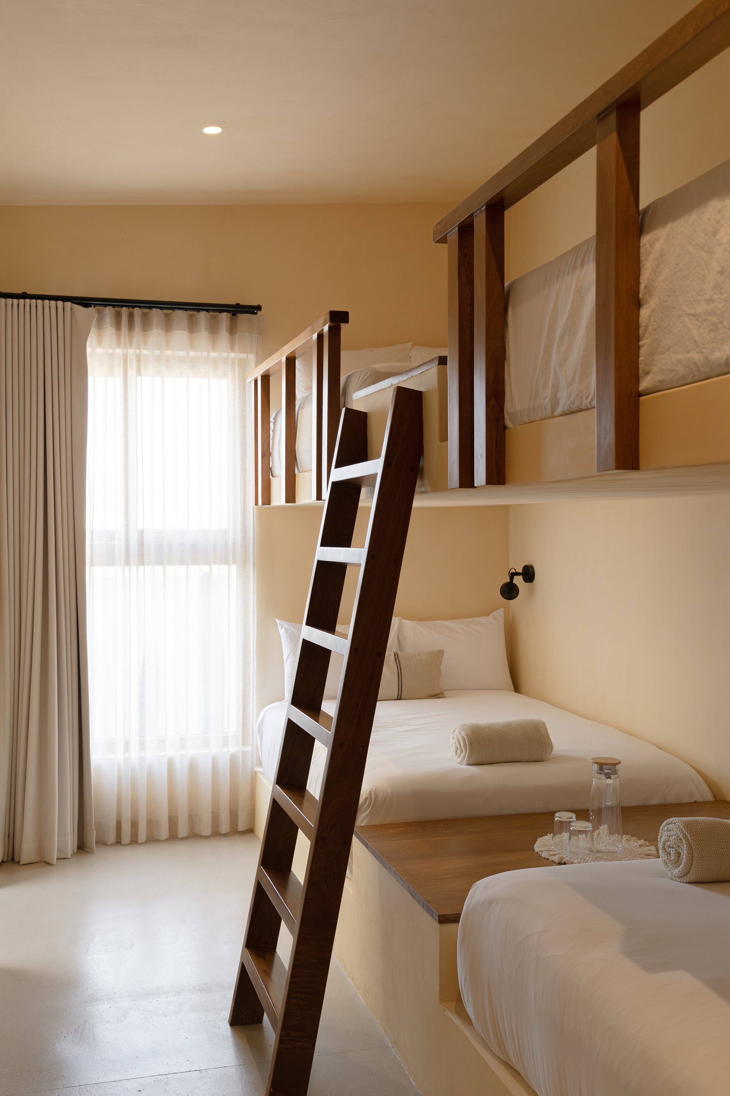 Minimalist bedroom with a lofted bunk bed, wooden ladder, and natural light coming through sheer curtains.