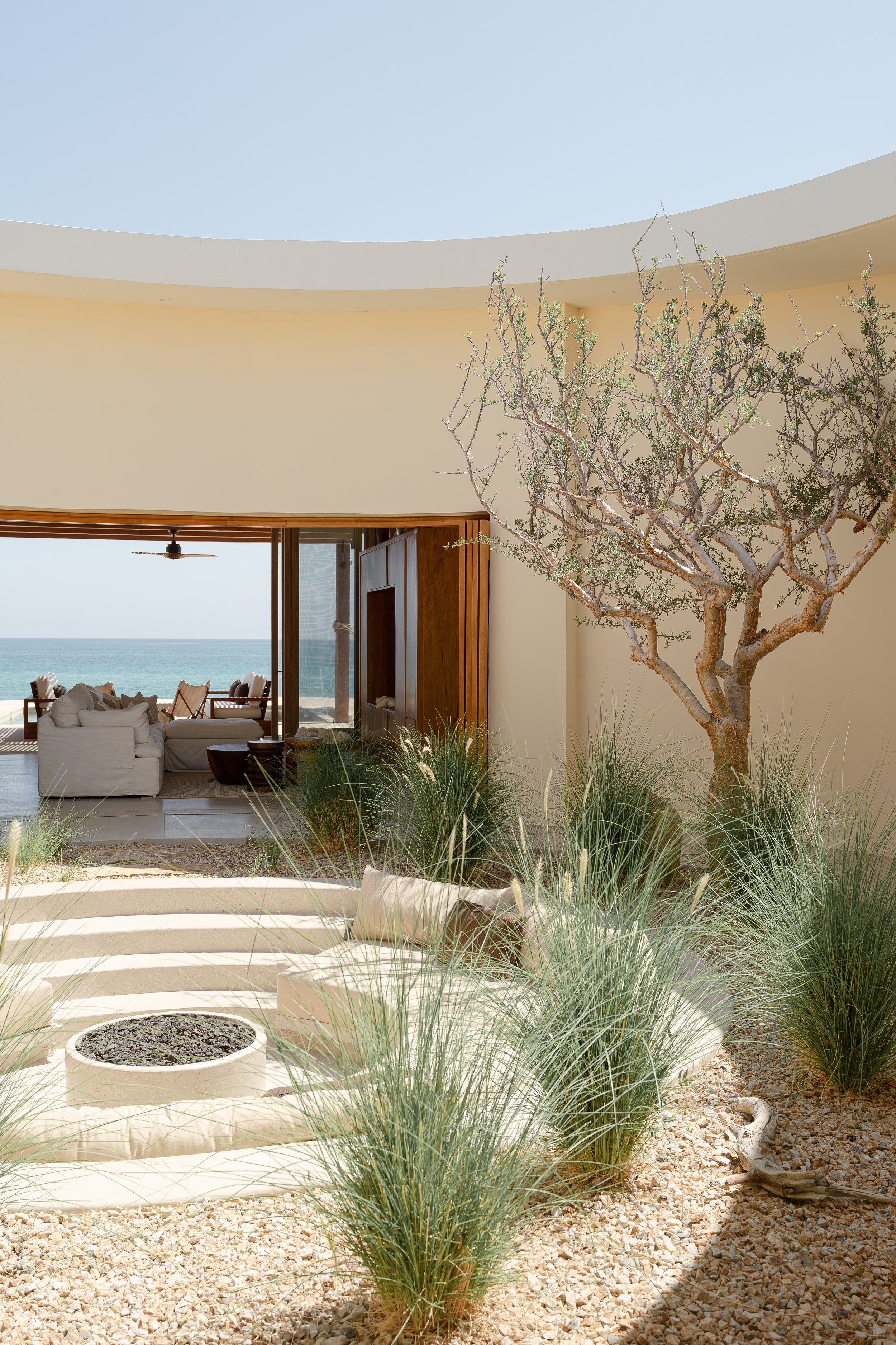 Modern outdoor patio area with circular seating, fire pit, decorative plants, and view of the ocean; minimalist architectural design with open space and natural elements.