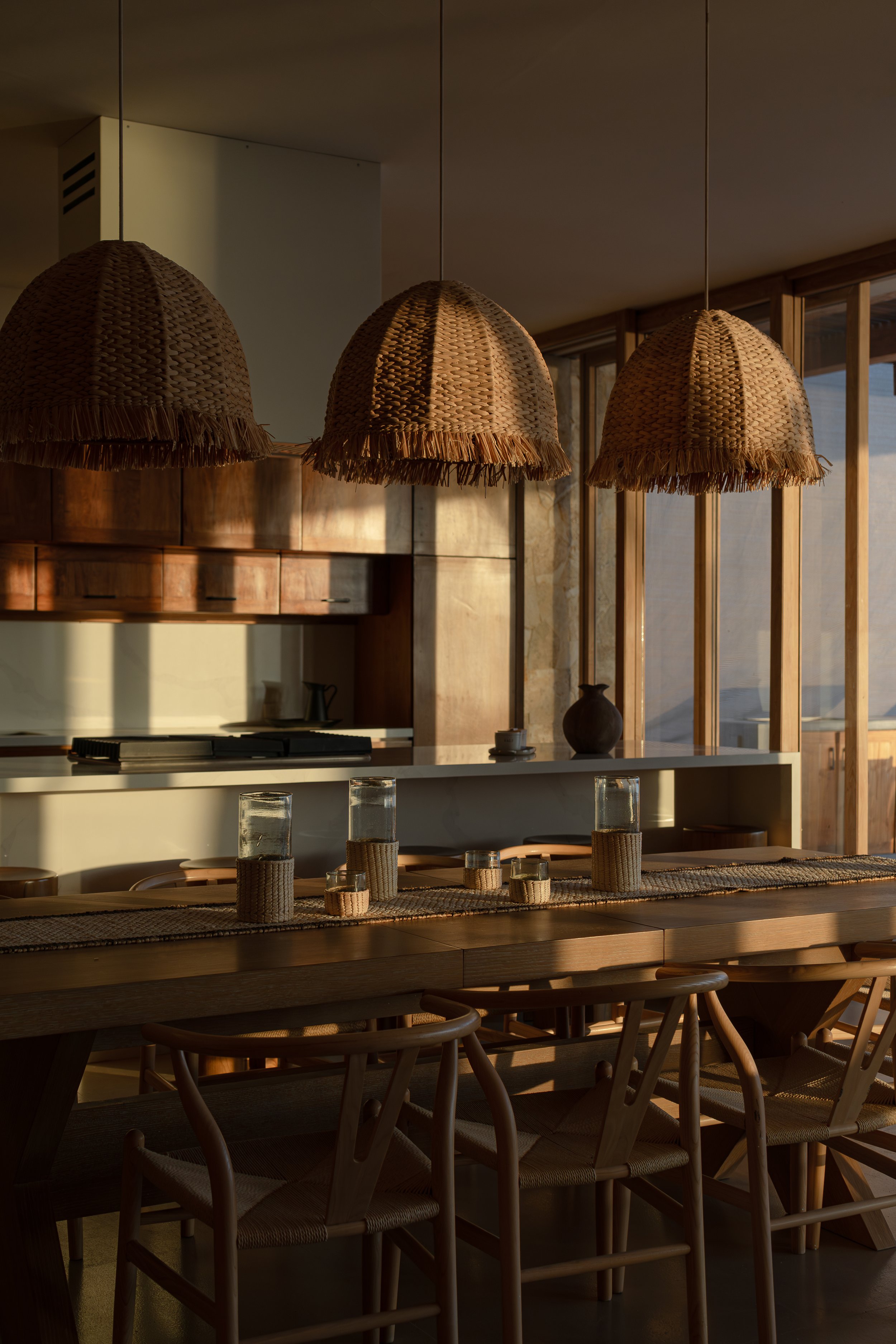 A stylish dining room with woven pendant lights hanging above a large wooden table set with woven placemats and glass containers. The room has wooden cabinetry and large windows allowing natural light to enter.