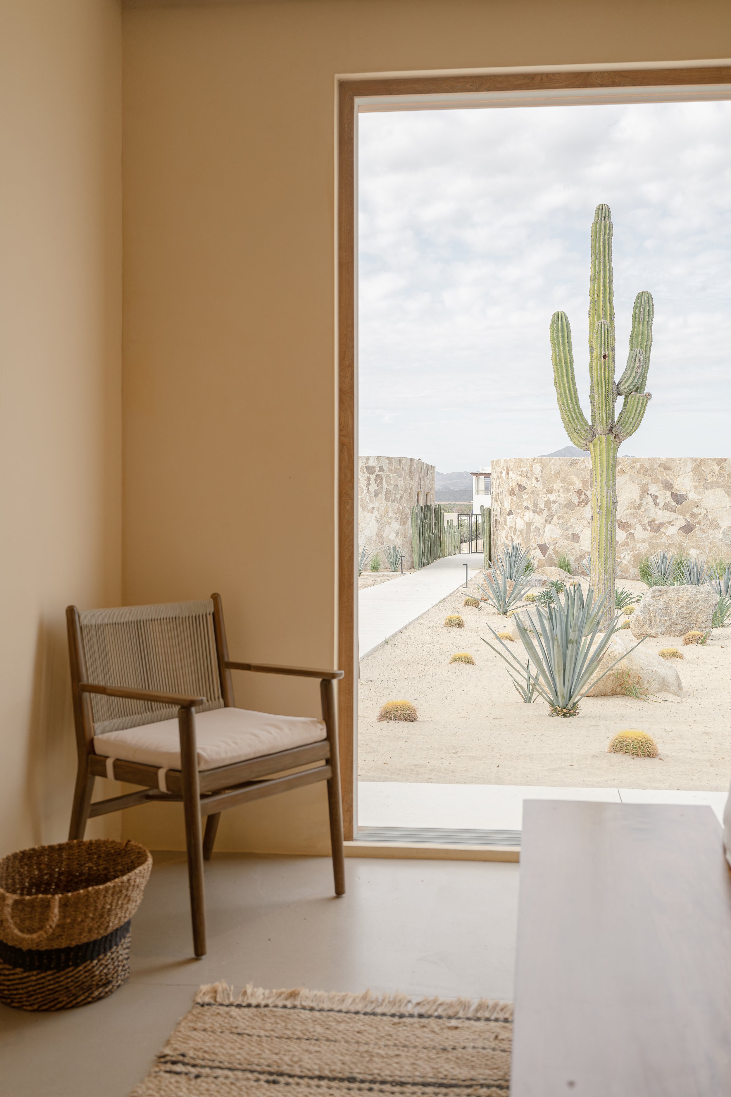 Interior room with beige walls, wooden chair with cushion, woven basket, and view of outdoor desert landscape with tall cactus, stone wall, and sandy pathway.