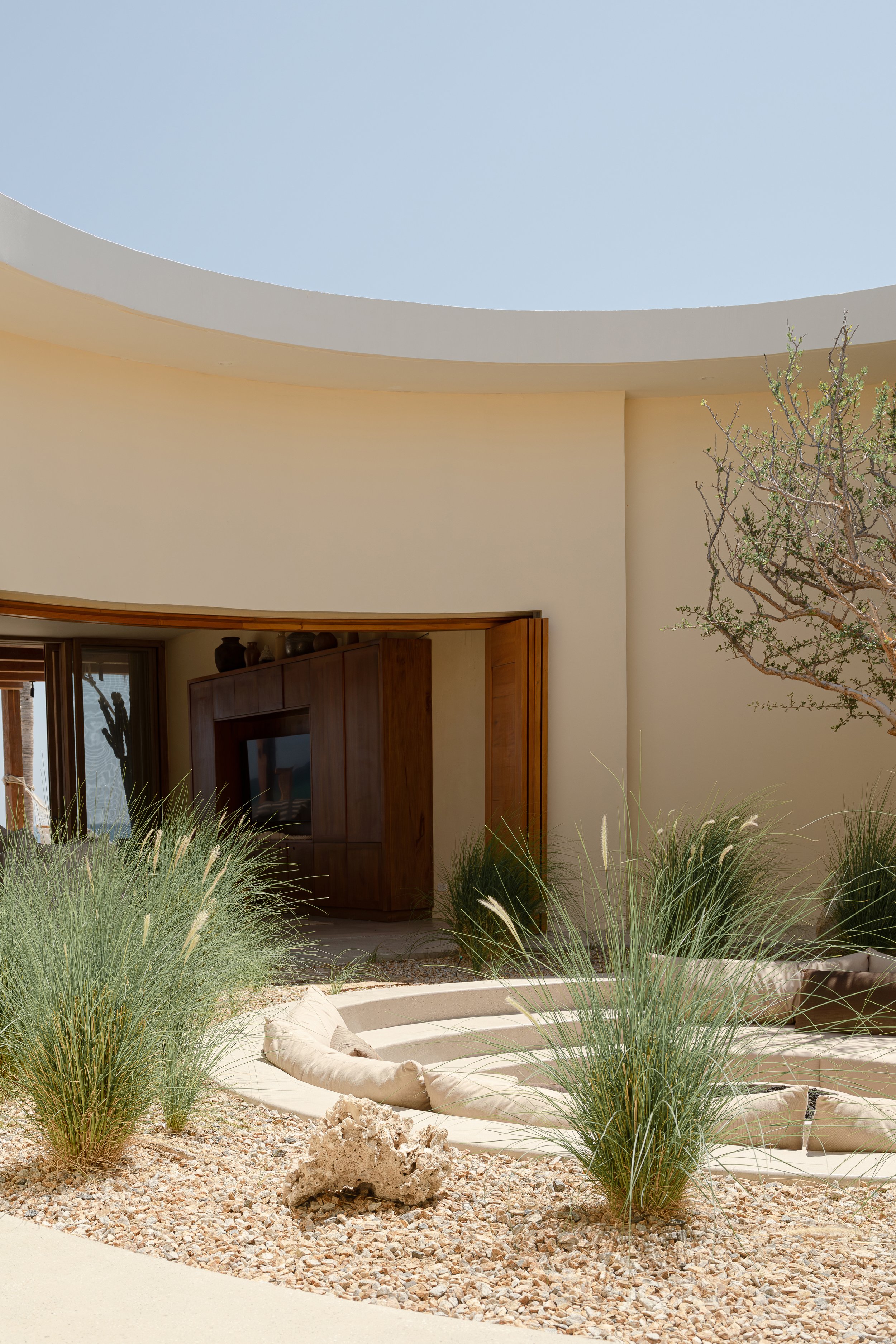 Modern outdoor patio with beige walls, a curved seating area with cushions, desert plants, and rocks under a clear blue sky.