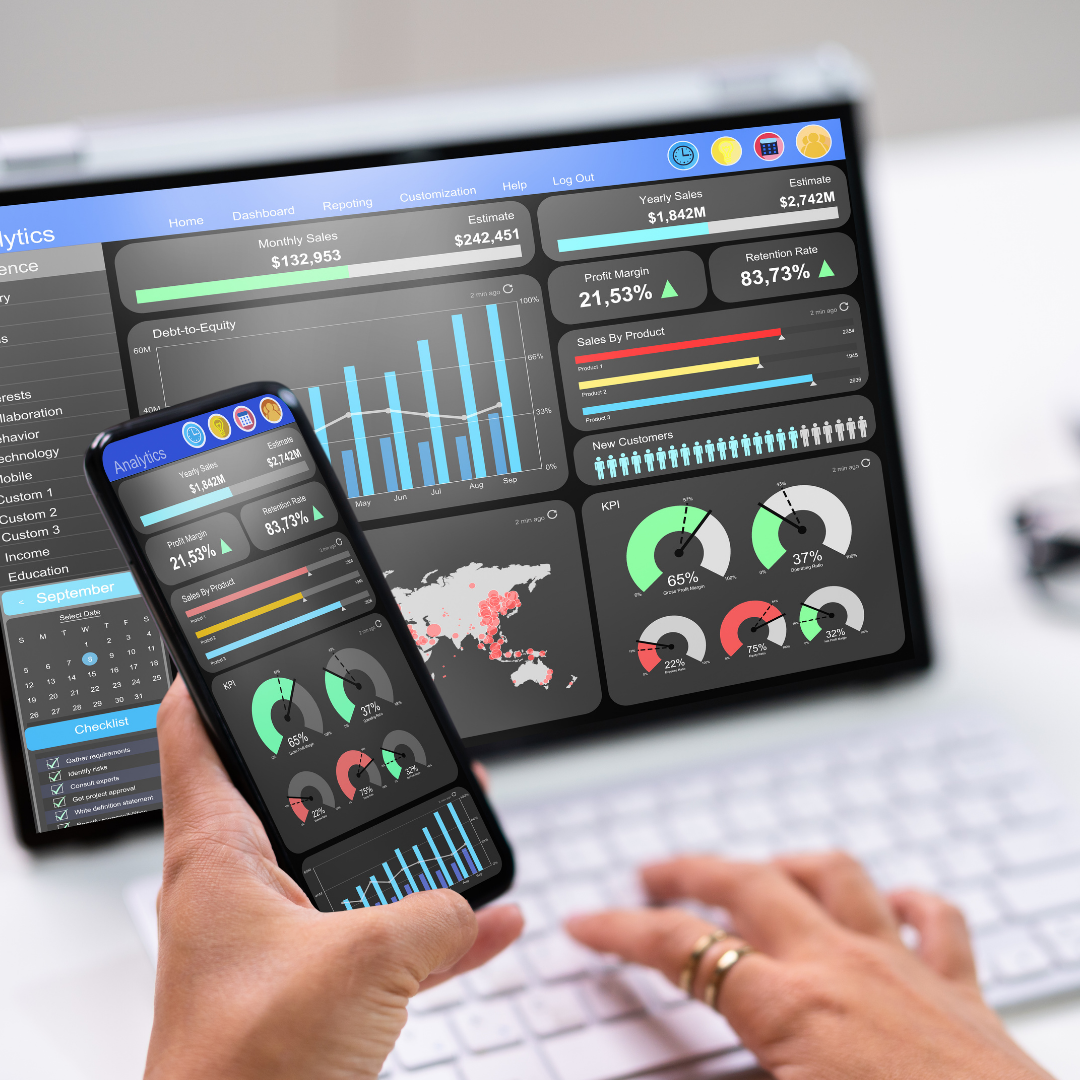 Person using a smartphone displaying business analytics graphs and charts, with a laptop screen showing similar analytics in the background.
