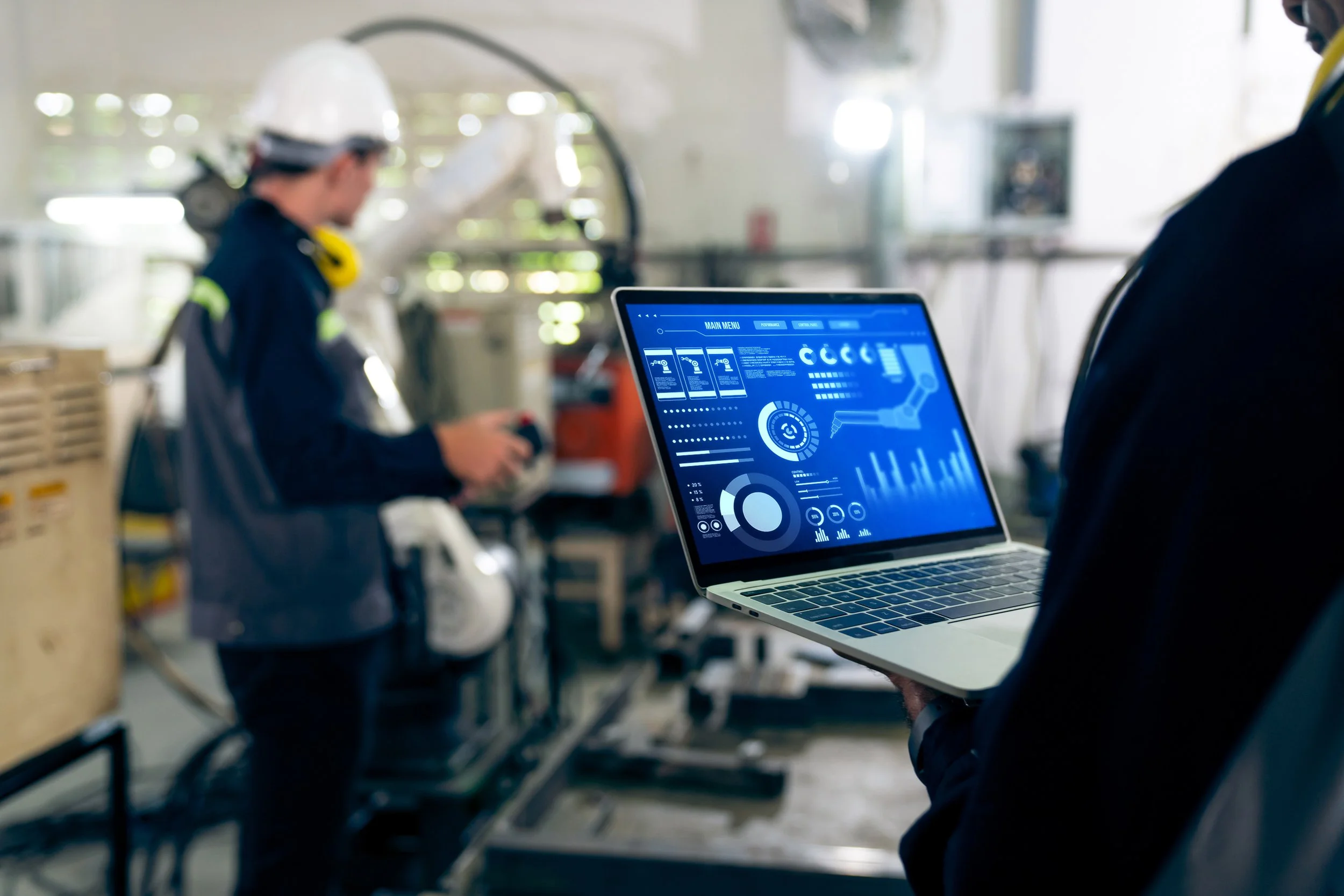 Person holding a laptop displaying data and charts, while a worker operates machinery in an industrial setting.