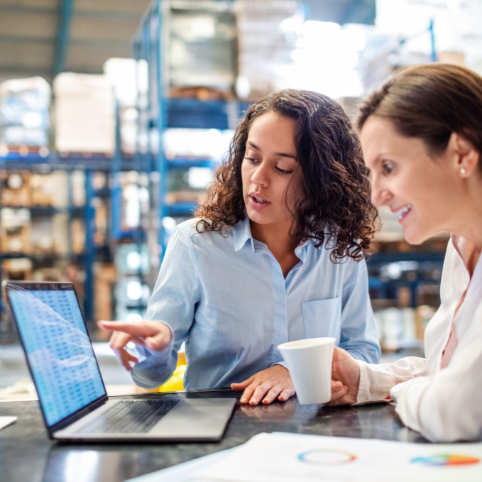 Two women discussing data on a laptop in a warehouse, one pointing at the screen, the other holding a coffee mug.