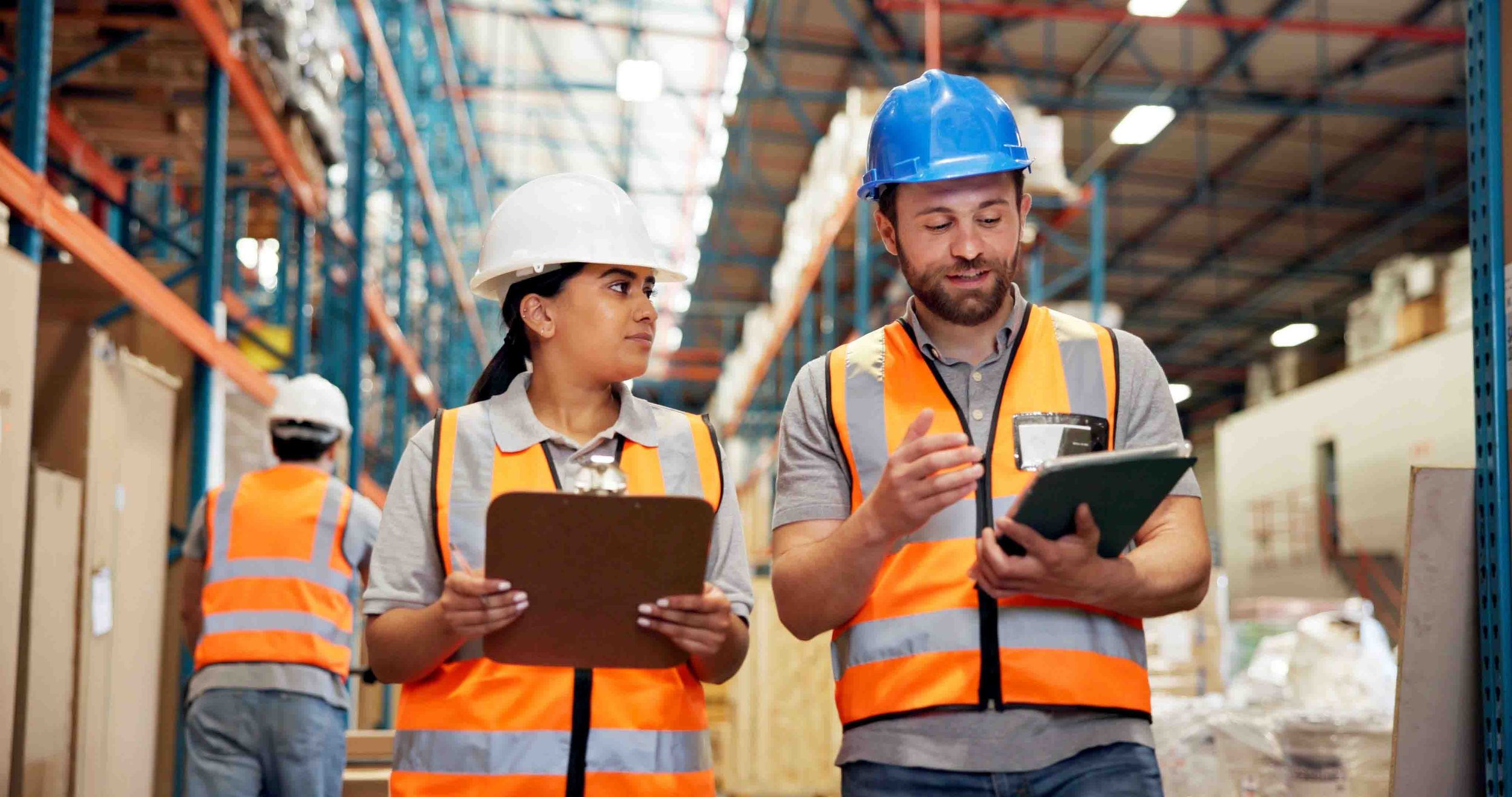 Two warehouse workers, a woman and a man, wearing safety helmets and reflective vests, stand in a large storage area. The woman holds a clipboard, and the man is holding a tablet and pointing at it, discussing something. In the background, another worker is seen wearing similar safety gear.