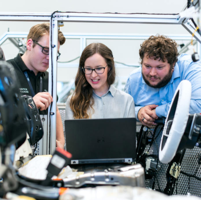 Three students in a high-tech lab gathered around a laptop, looking at it with interest and smiling, surrounded by robotic equipment and machinery.