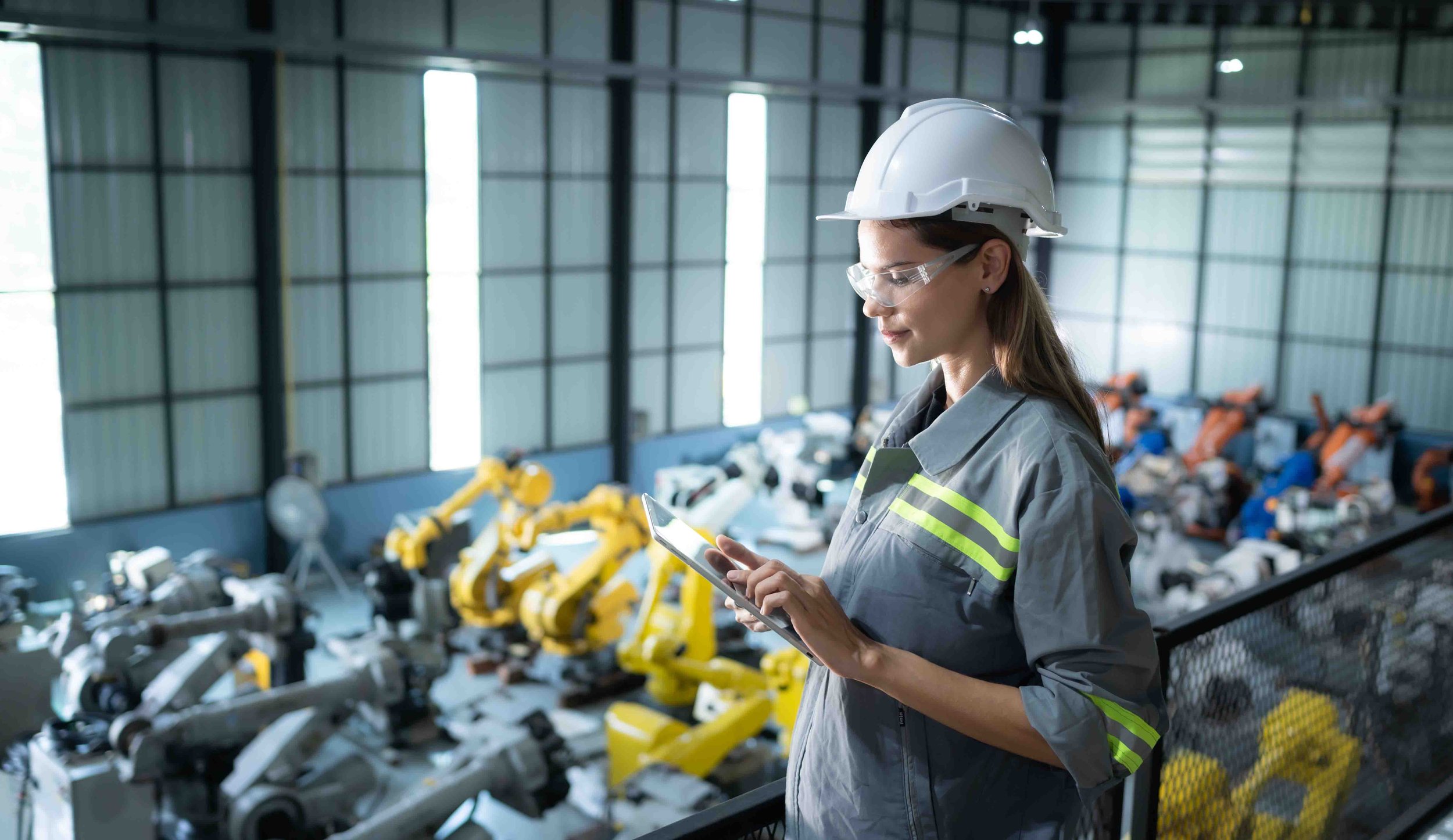 A female engineer wearing safety goggles and a hard hat inspecting machinery inside an industrial warehouse using a tablet.
