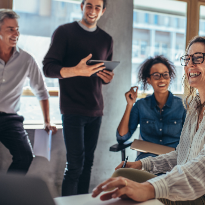 Four colleagues in a bright office smiling and laughing during a meeting or discussion, with one standing and holding a tablet, others sitting with notebooks and a laptop.