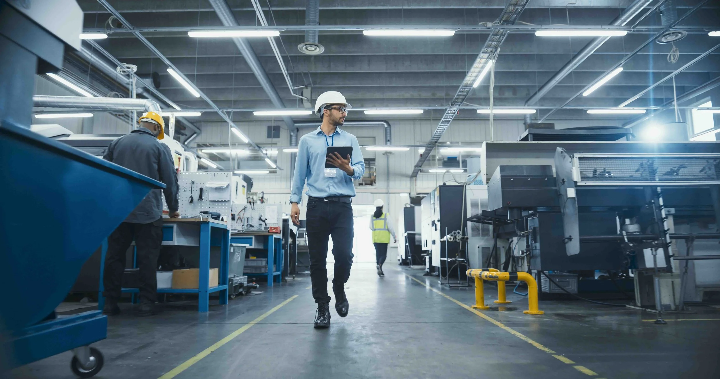 A man in business attire with a safety helmet and tablet walking through an industrial manufacturing facility with workers and machinery.