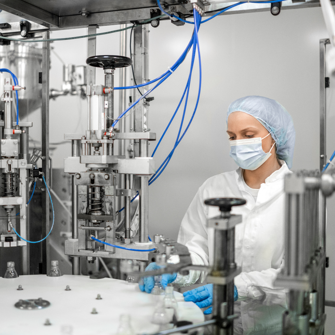 A scientist wearing a mask, hair cover, and gloves working on a manufacturing machine with bottles in a clean industrial setting.