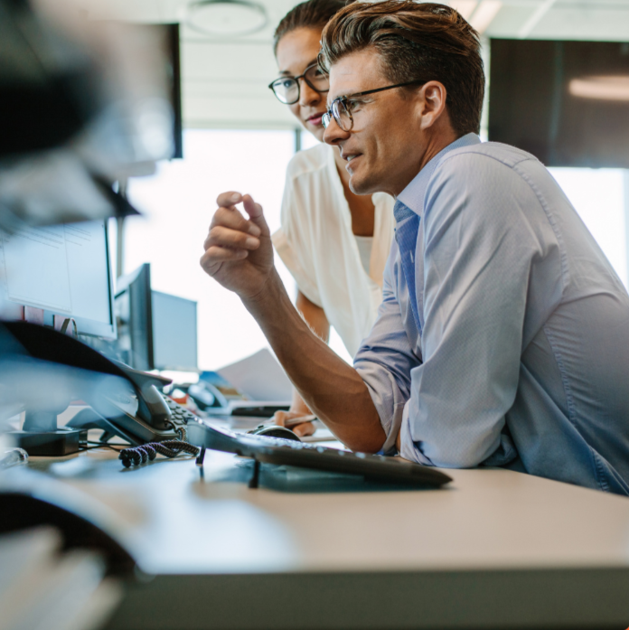 Two professionals discussing at a desk with multiple computer monitors in an office.