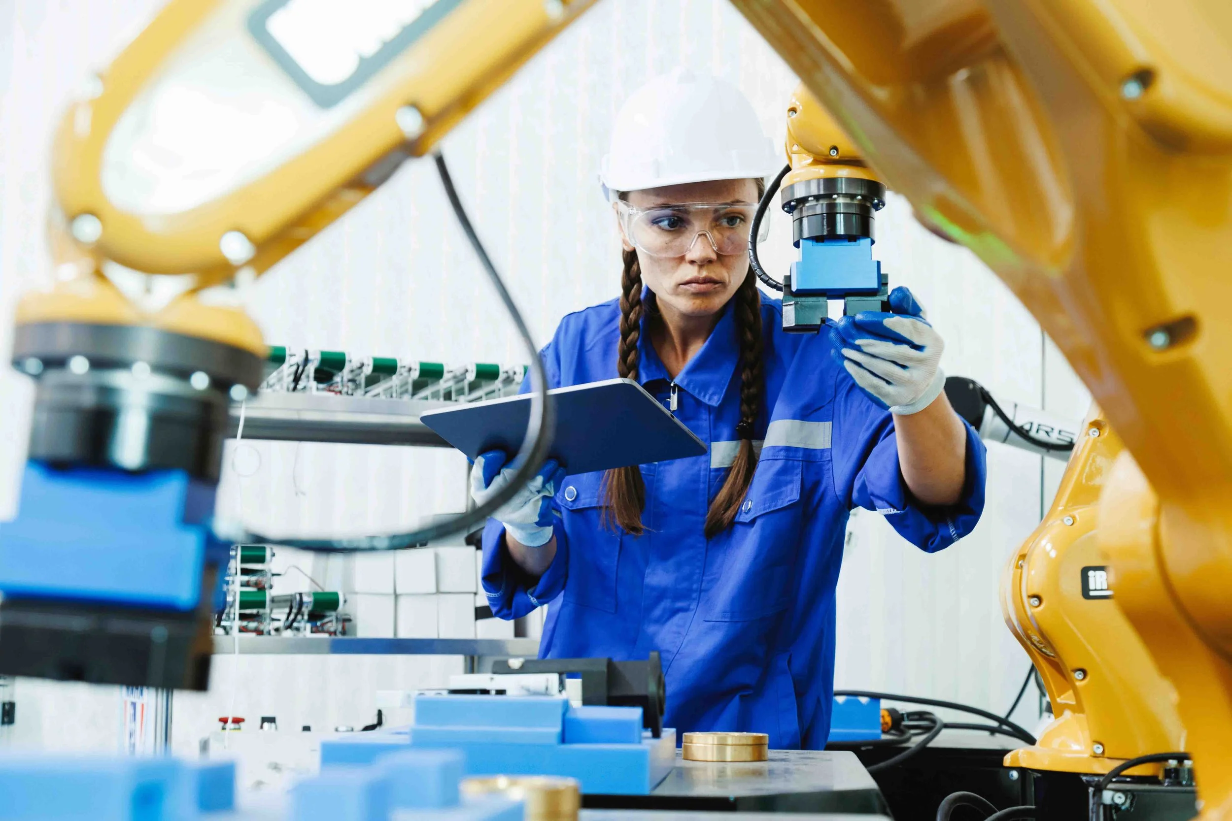 A female engineer in safety glasses and gloves operating a robotic arm with a microscope in an industrial setting, holding a clipboard.