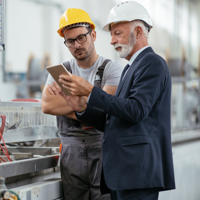 Two men, one young and one older, wearing safety helmets and inspecting equipment in an industrial setting.
