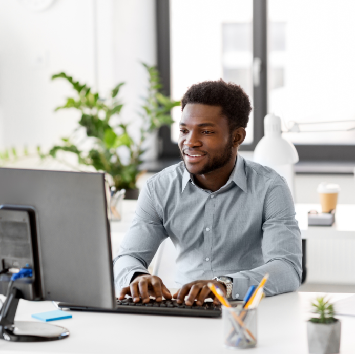 A young man working on a desktop computer in a bright, modern office with a large window, green plants, and office supplies on the desk.