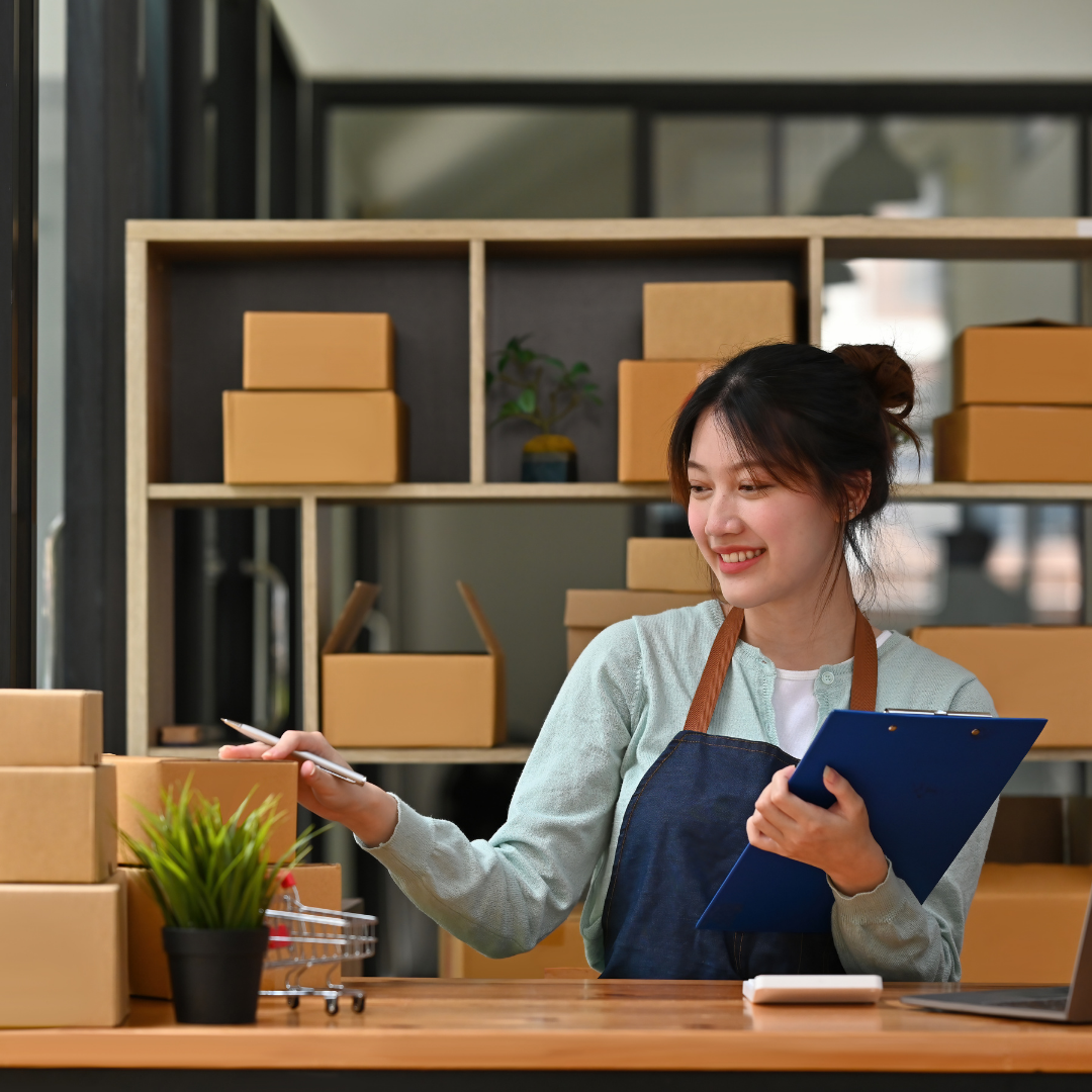 A young woman in an apron smiling while working with packages and a notepad at a store or warehouse.
