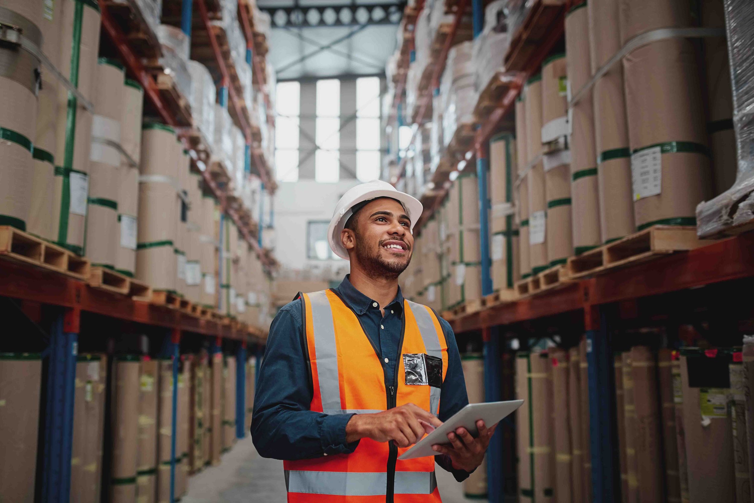 A man in safety vest and hard hat holding a tablet, standing in a warehouse aisle with shelves filled with large boxes using fuuz an industrial intelligence platform for inventory tracking