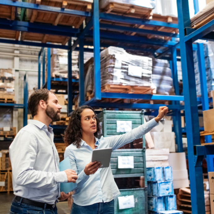 Two warehouse workers, a man and a woman, discussing inventory in a storage facility. The woman is holding a tablet and pointing at stacked shelves with boxes and pallets.