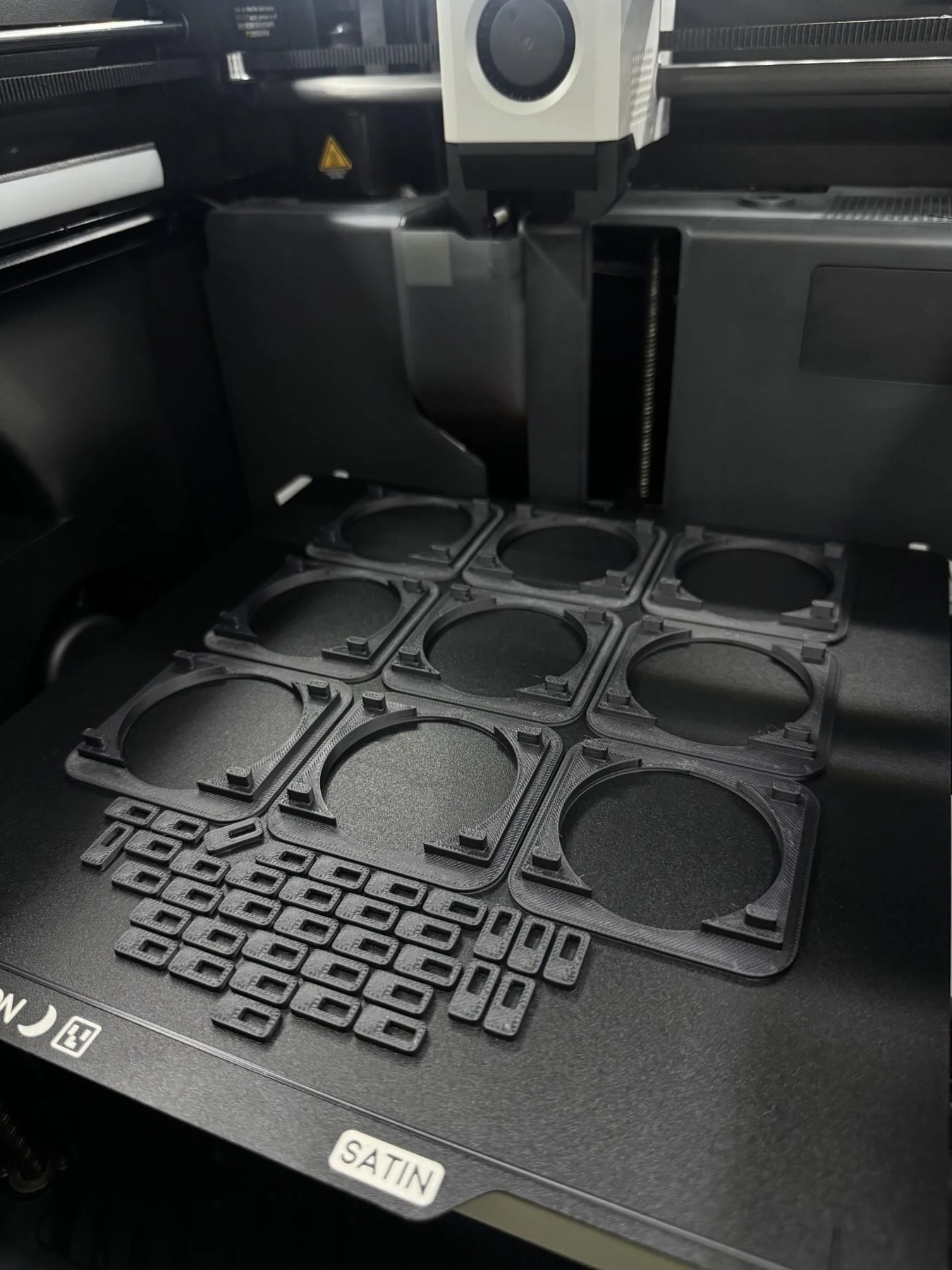 The image shows the interior of a 3D printer, with various black plastic parts and frames, likely for assembling or printing objects. The printer has a black and white print head and a dark surface with labels indicating material options like 'SATIN'.