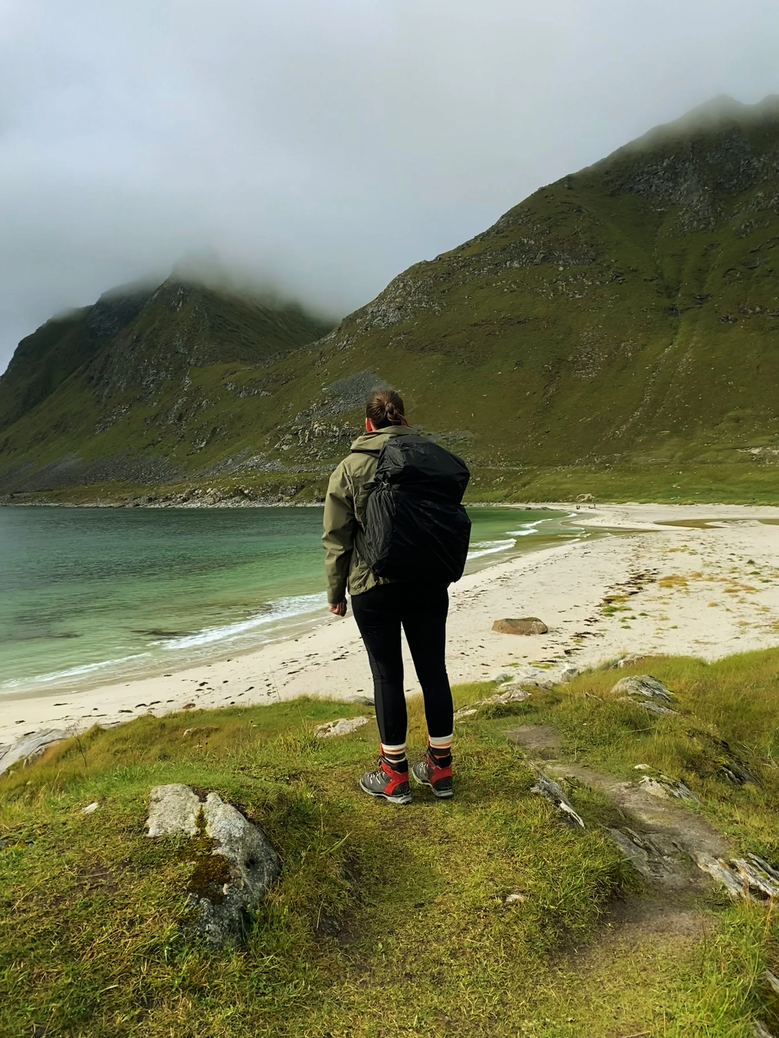 Eine Person steht an einem Strand mit grünen Bergen im Hintergrund. Es ist bewölkt und die Person trägt Wanderausrüstung mit Rucksack.
