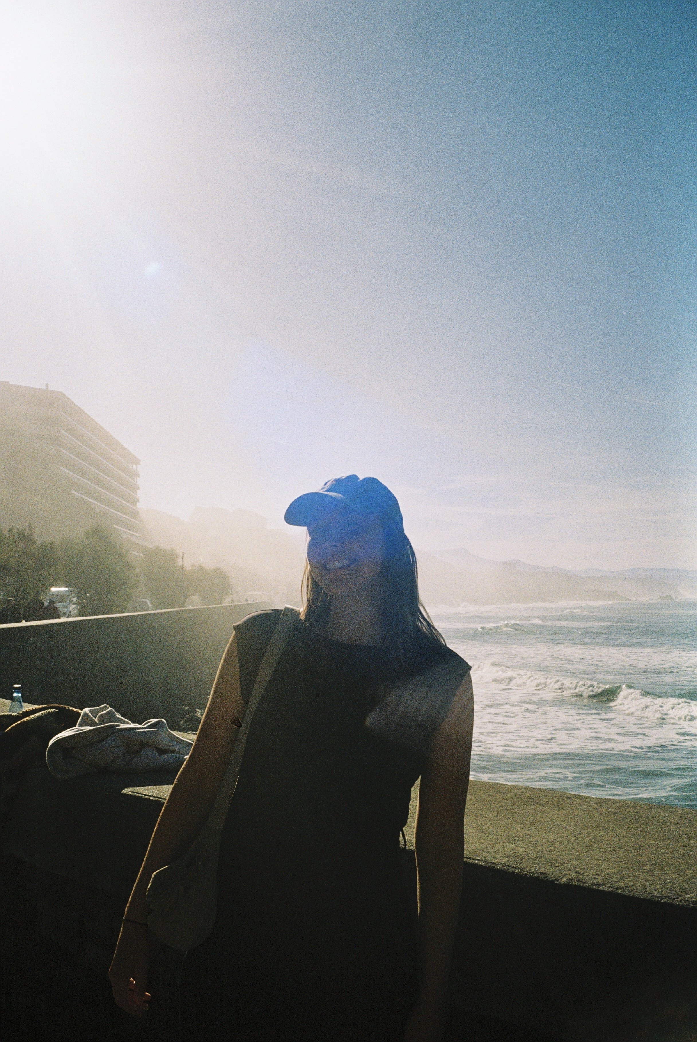 Eine Frau steht am Meer, trägt eine schwarze Kleidung, eine schwarze Mütze und trägt eine Tasche. Die Sonne scheint stark, und es gibt eine Küstenlandschaft im Hintergrund.