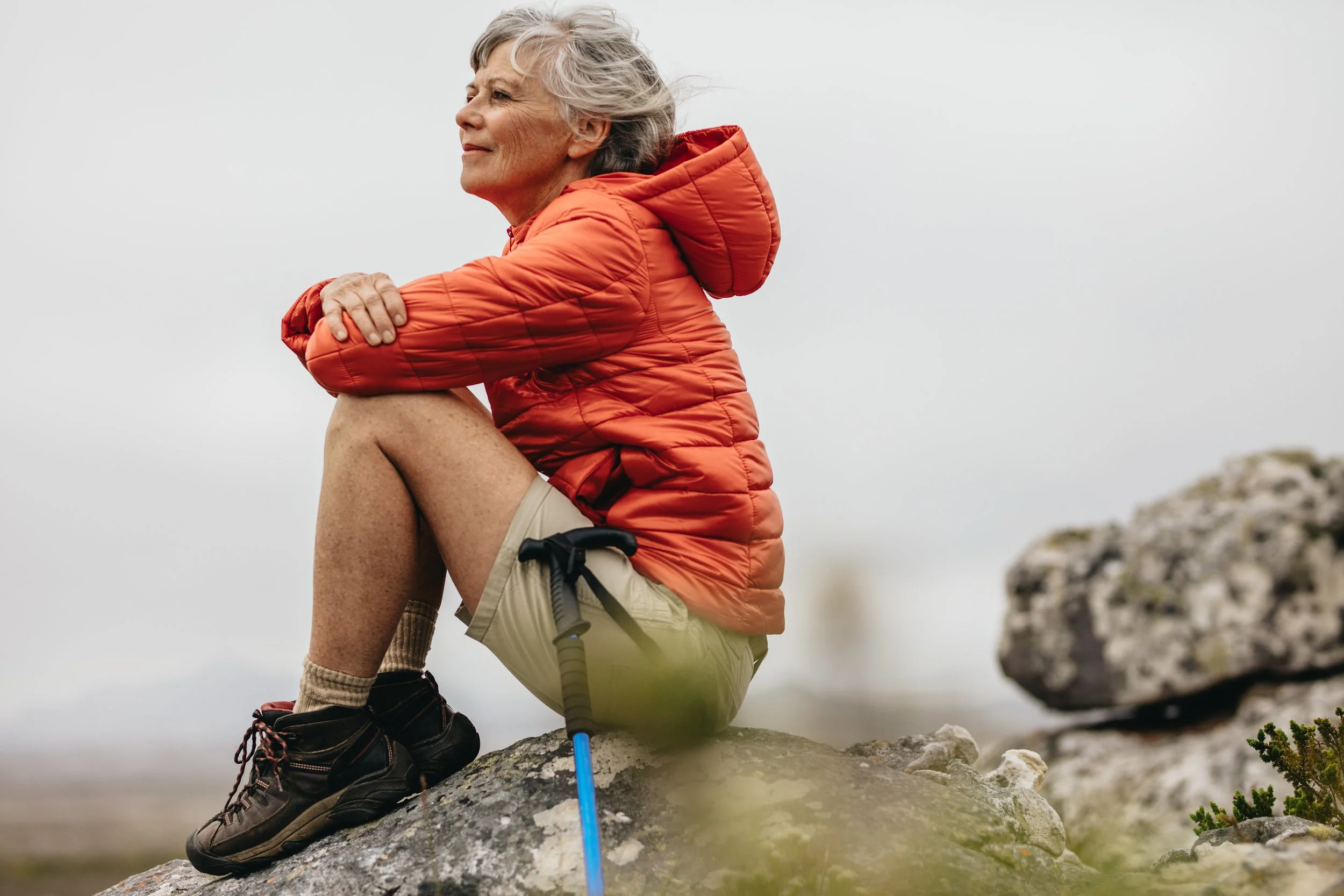 Senior woman in red jacket sitting on a rock during a hike, holding trekking poles, looking thoughtful.