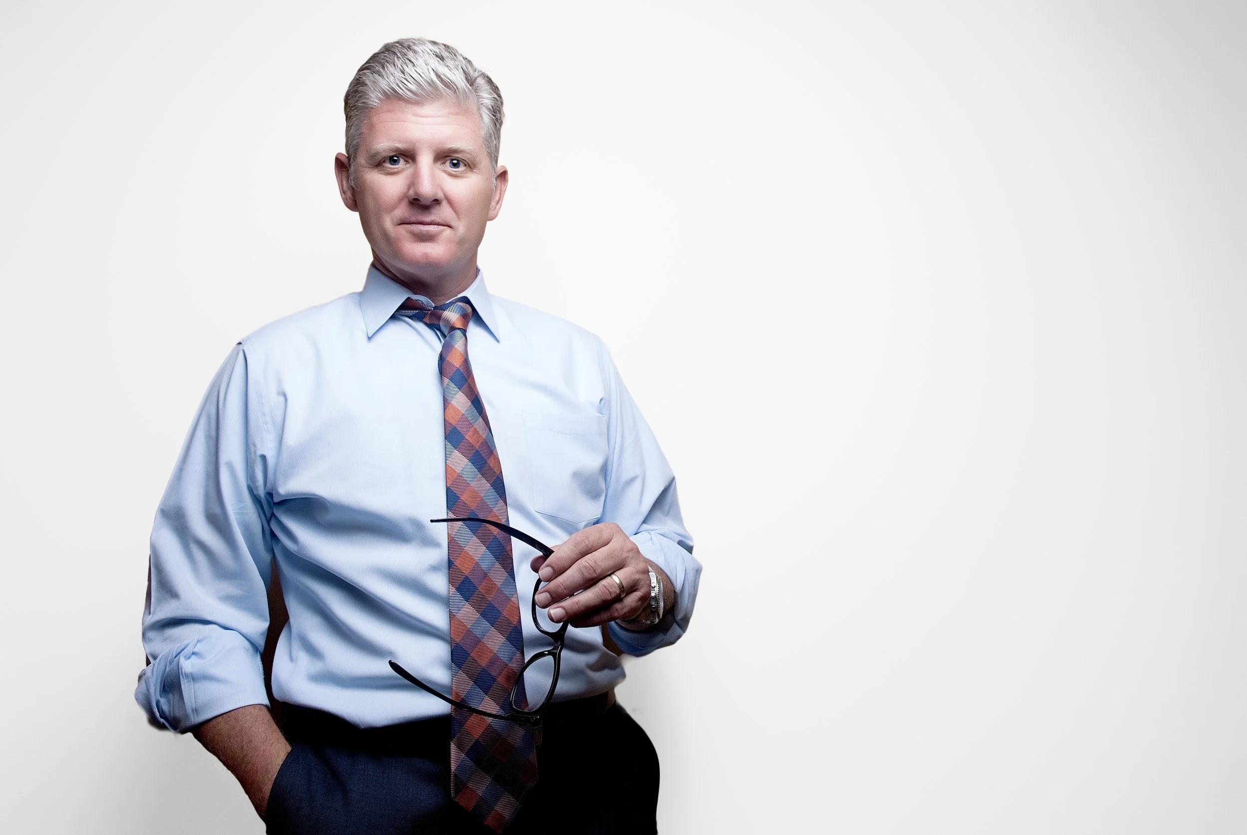 A middle-aged man with gray hair, wearing a light blue dress shirt, a colorful plaid tie, and black trousers, holding eyeglasses in his right hand and standing against a plain white background.