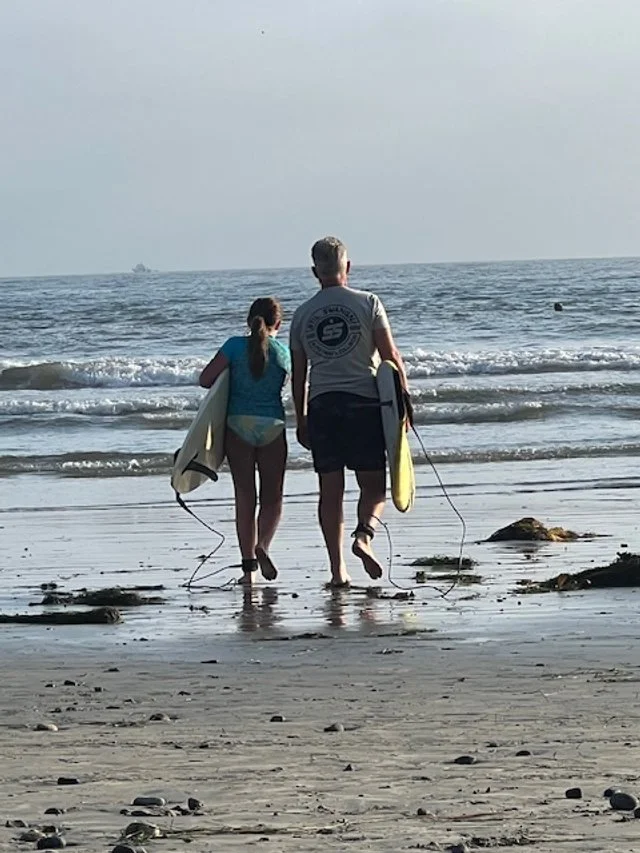 A man and a girl walking on a beach carrying surfboards towards the ocean.