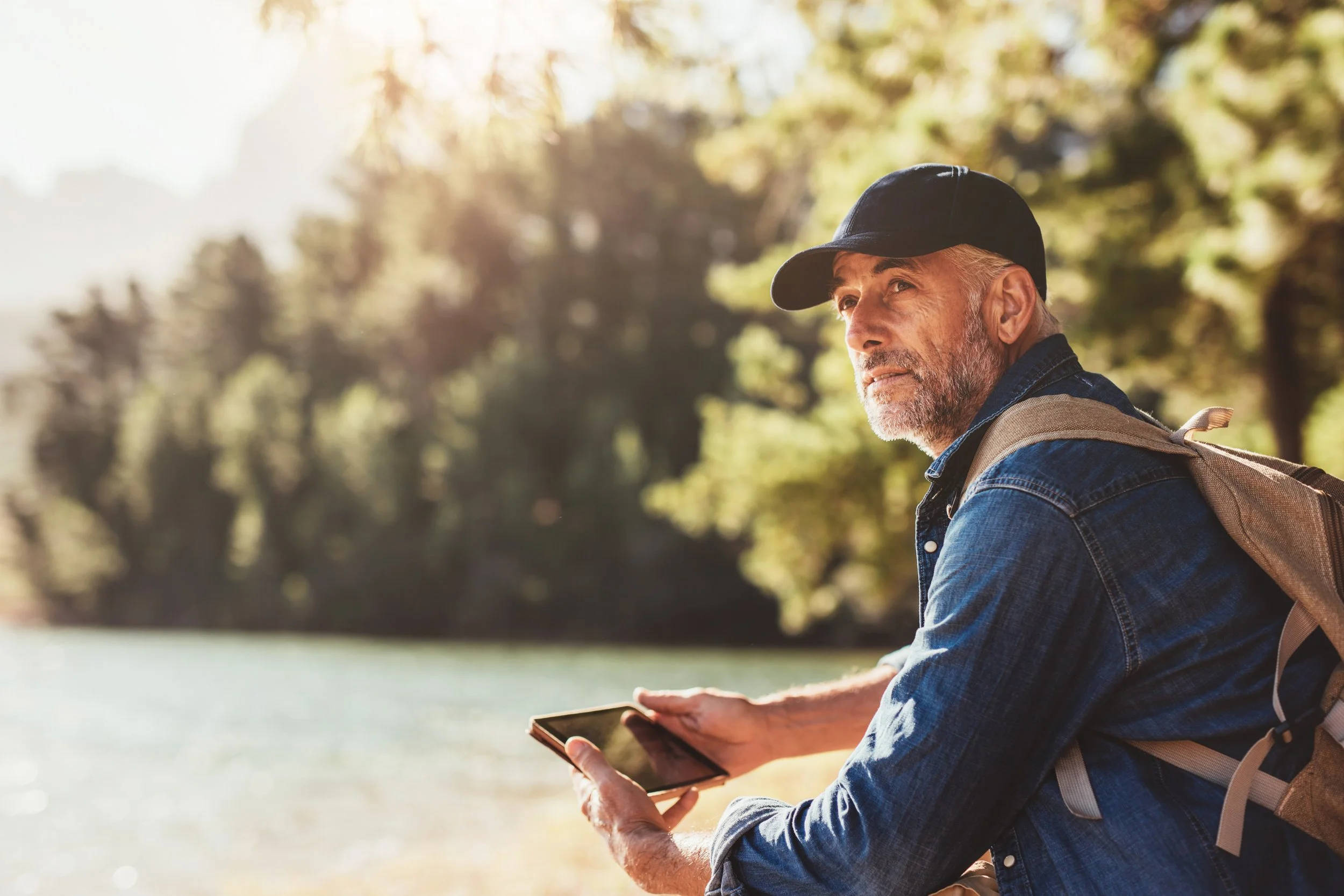 Middle-aged man in denim jacket and cap holding a tablet outside by a lake during the day.