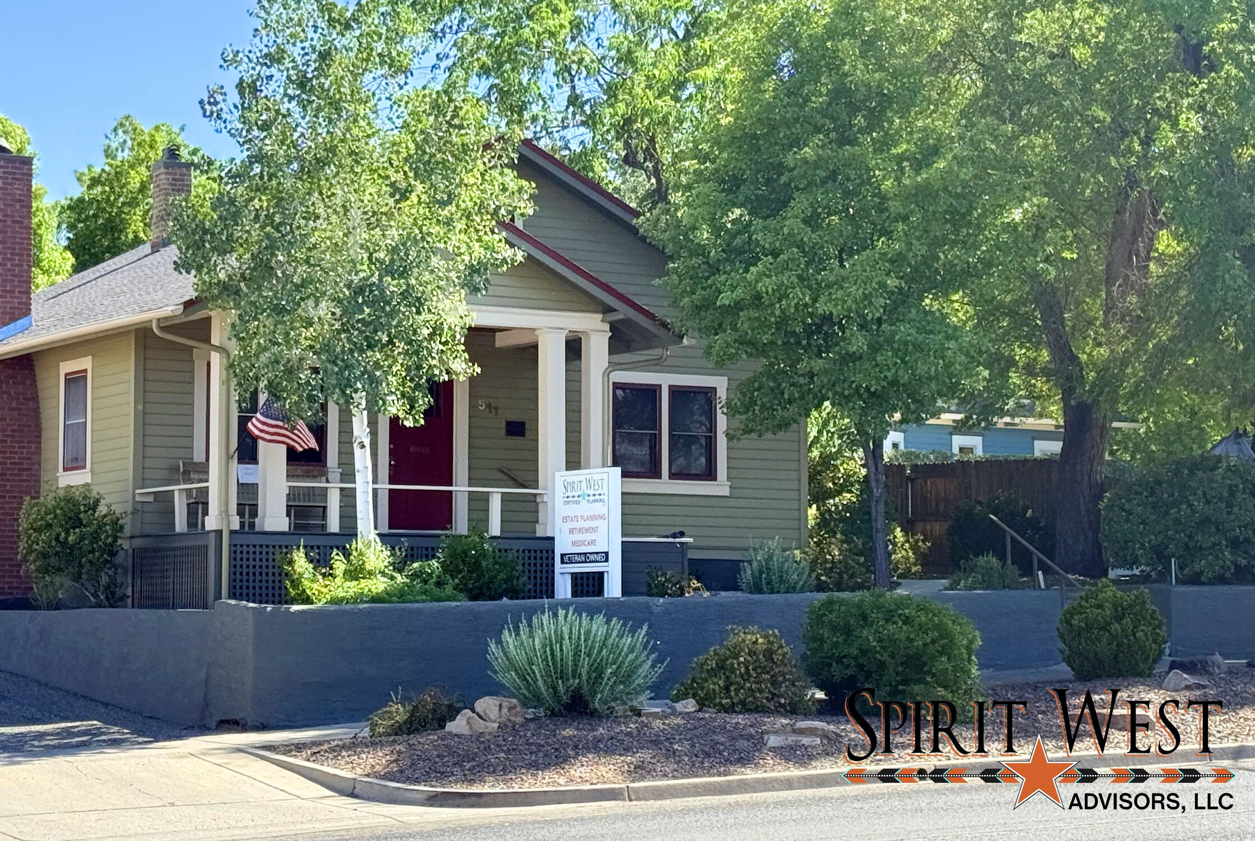 A light green house with dark red door and window trim, a porch with white railings, an American flag on the porch, surrounded by trees and bushes, situated above a sidewalk and street near a gray retaining wall. A sign on the porch reads: 'Spirit West Advisors, LLC'.
