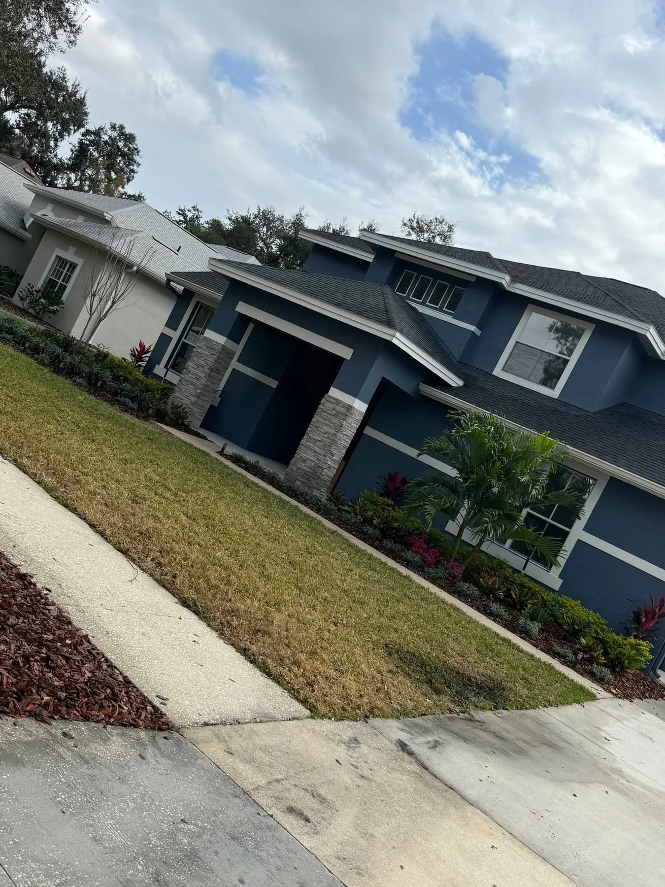 A modern blue house with white trim, stone accents, and a dark roof, surrounded by landscaped yard with grass, small palm tree, and flowering plants, under a partly cloudy sky. Beautiful manicured landscaping custom created by Lawn Care Plus LLC.