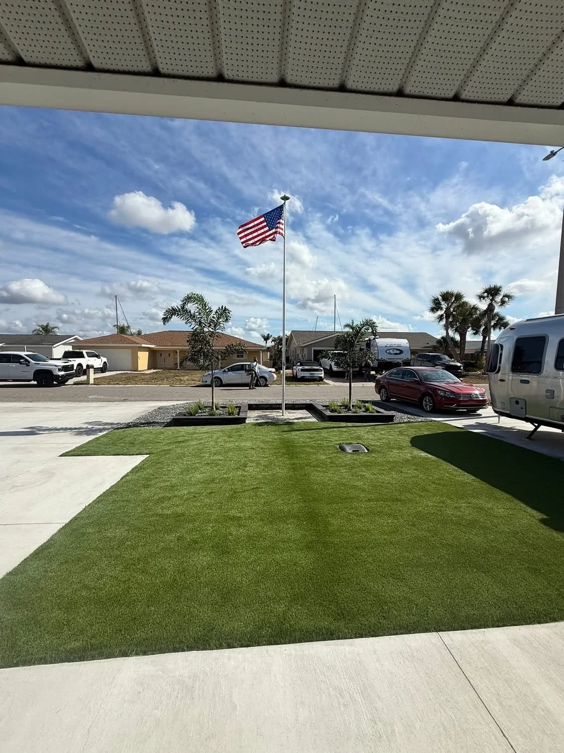 American flag flying on a flagpole in a neighborhood with houses, palm trees, and parked cars under a partly cloudy sky. Beautiful manicured landscaping custom created by Lawn Care Plus LLC.