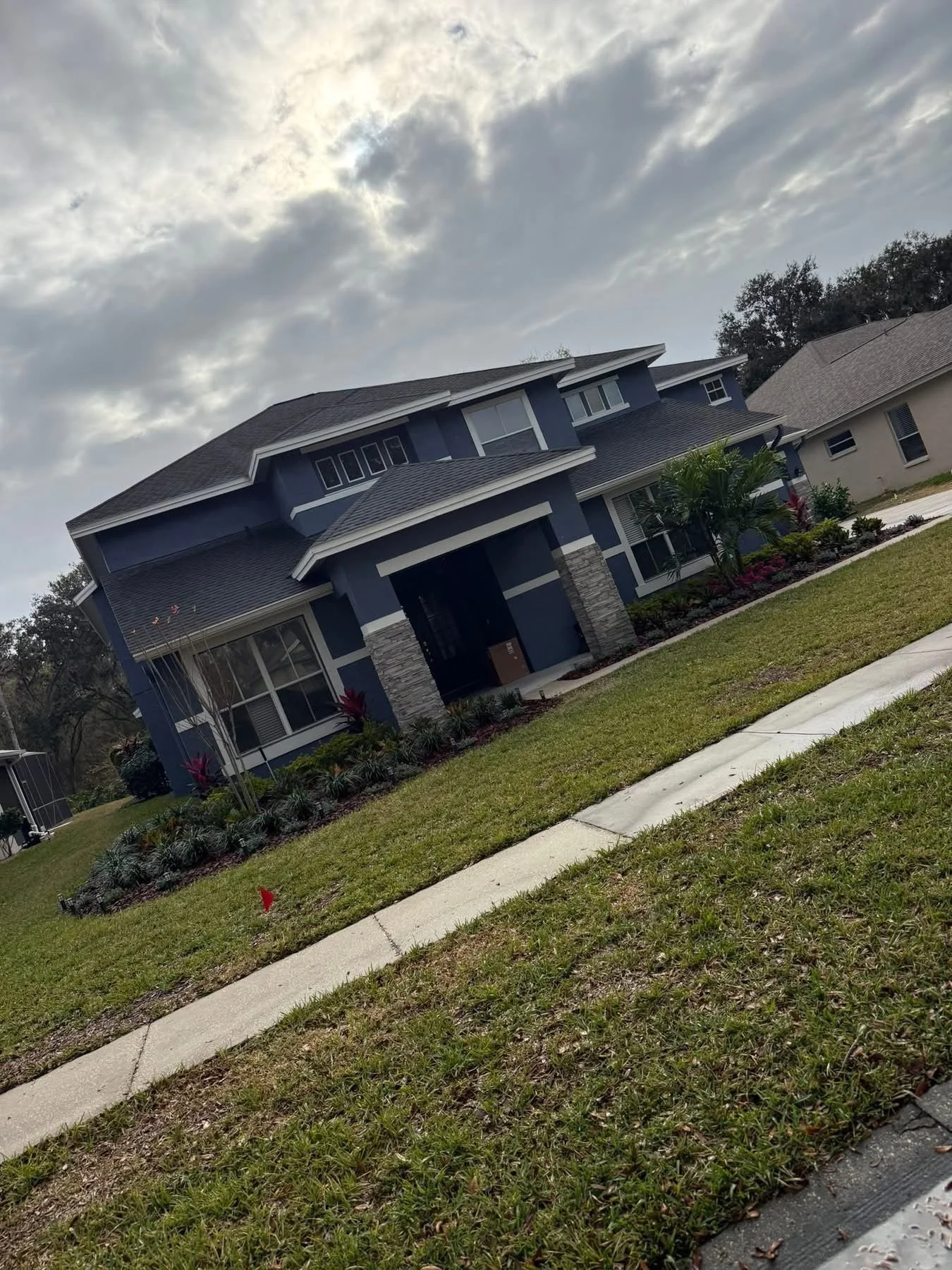 A modern two-story blue house with white trimming and stone accents on the front porch, surrounded by a green lawn, palm trees, and landscaped flower beds, under a cloudy sky. Beautiful manicured landscaping custom created by Lawn Care Plus LLC.