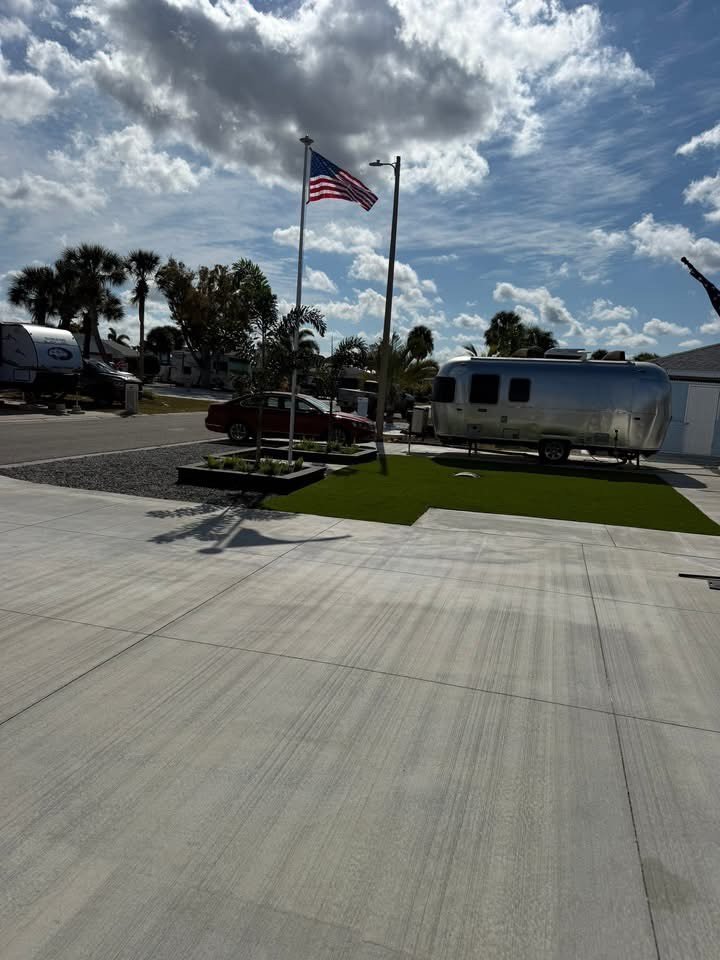 A flagpole with an American flag, parked trailers including an Airstream trailer, a red car, a few palm trees, and a partly cloudy sky. Beautiful manicured landscaping custom created by Lawn Care Plus LLC.