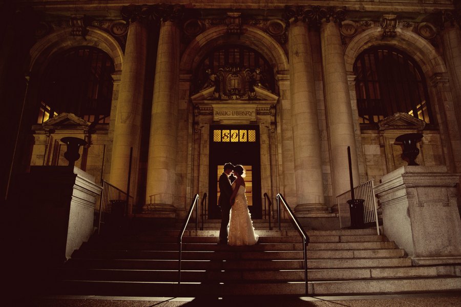 A couple in wedding attire stands close together on the steps of a grand, illuminated library at night, silhouetted against the building's entrance.