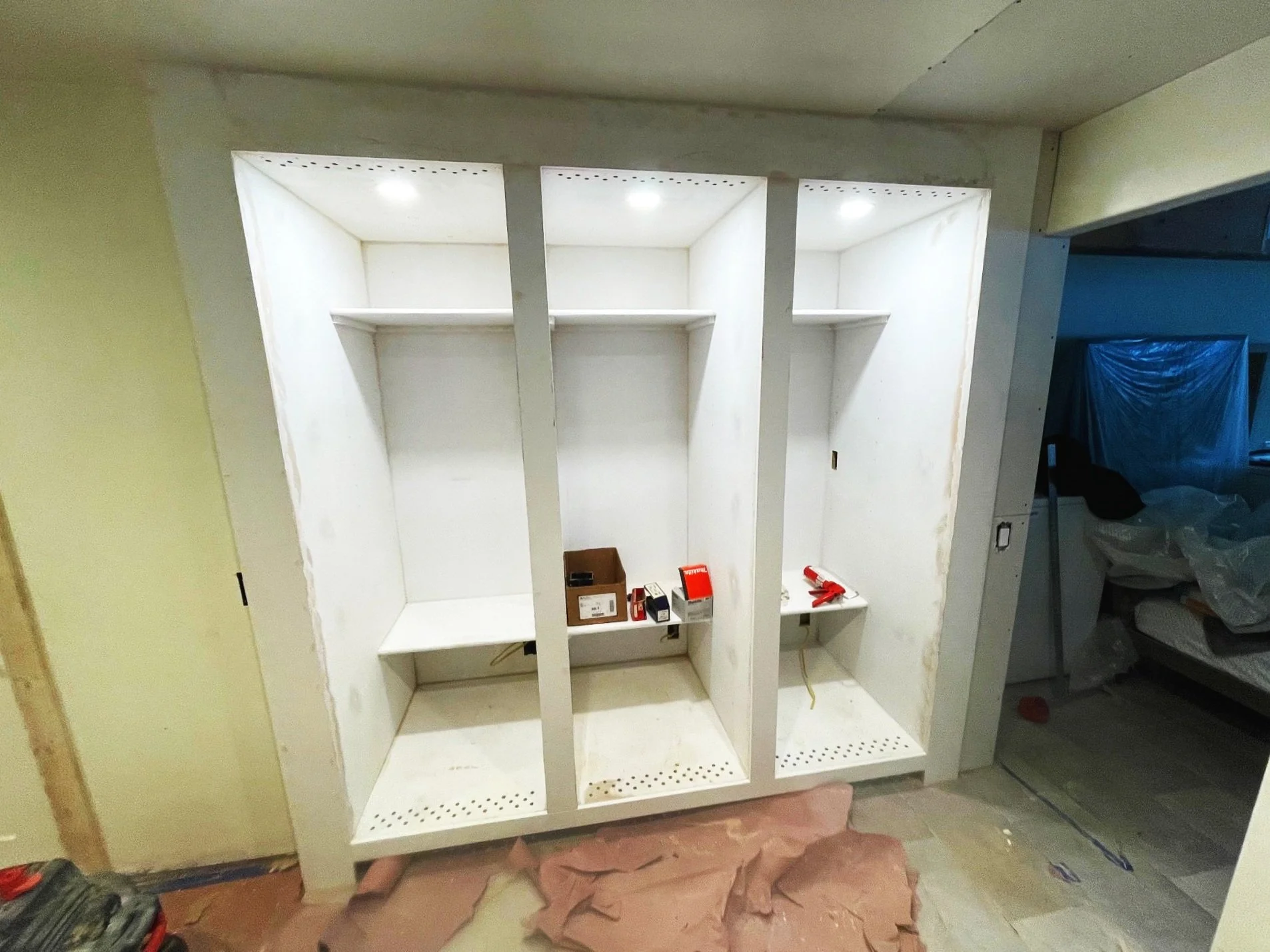 White built-in shelves or cabinets under construction with tools and supplies inside, in a room with unfinished walls and floor.