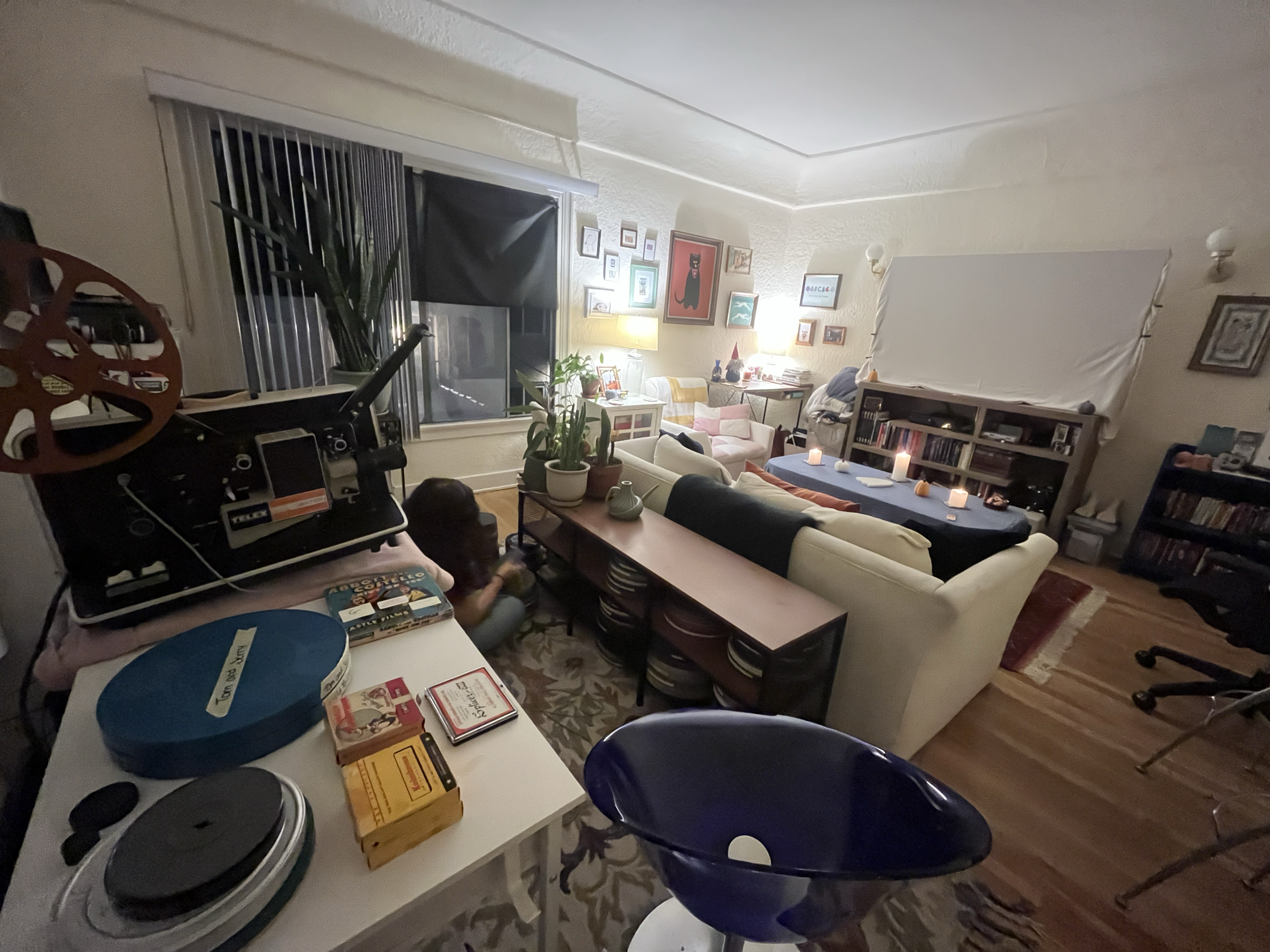 A cozy living room with a 16mm projector set up on the table, 16mm film reels on the table such as Tom and Jerry, Abbott and Costello, home movies and more. A projection screen is set up on the other side of the room.