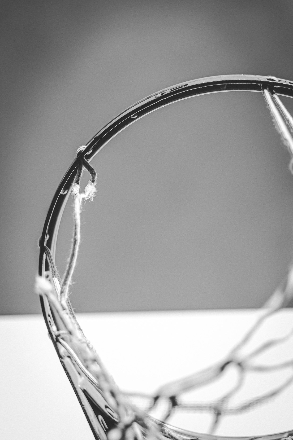 Close-up black and white photograph of a worn basketball net hoop with frayed strings and a blurred background.