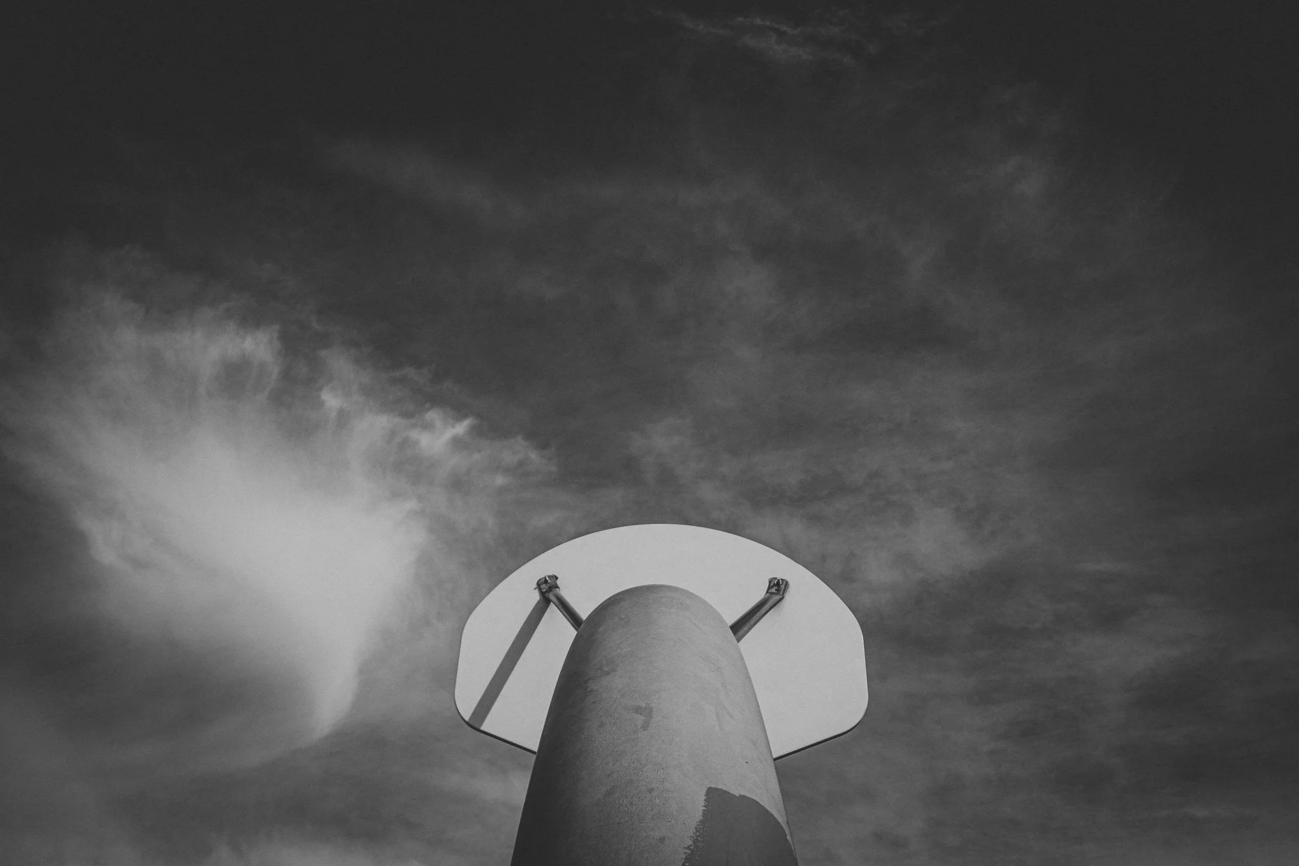 Black and white photo of a basketball hoop and backboard from below, with a cloudy sky in the background.