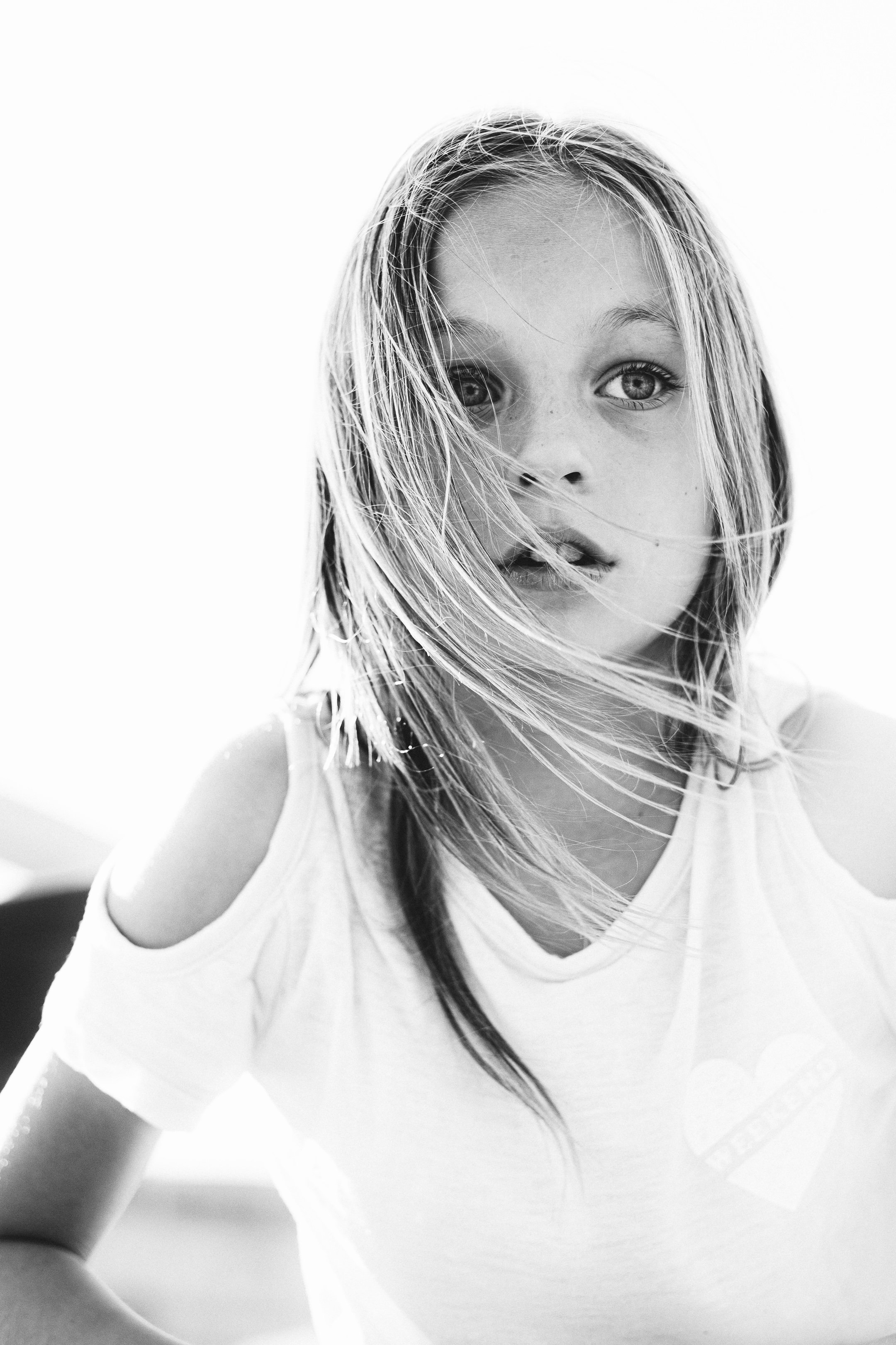 Close-up black and white photo of a young girl with wet hair, gazing directly at the camera, wearing a white T-shirt with cold shoulder cutouts.