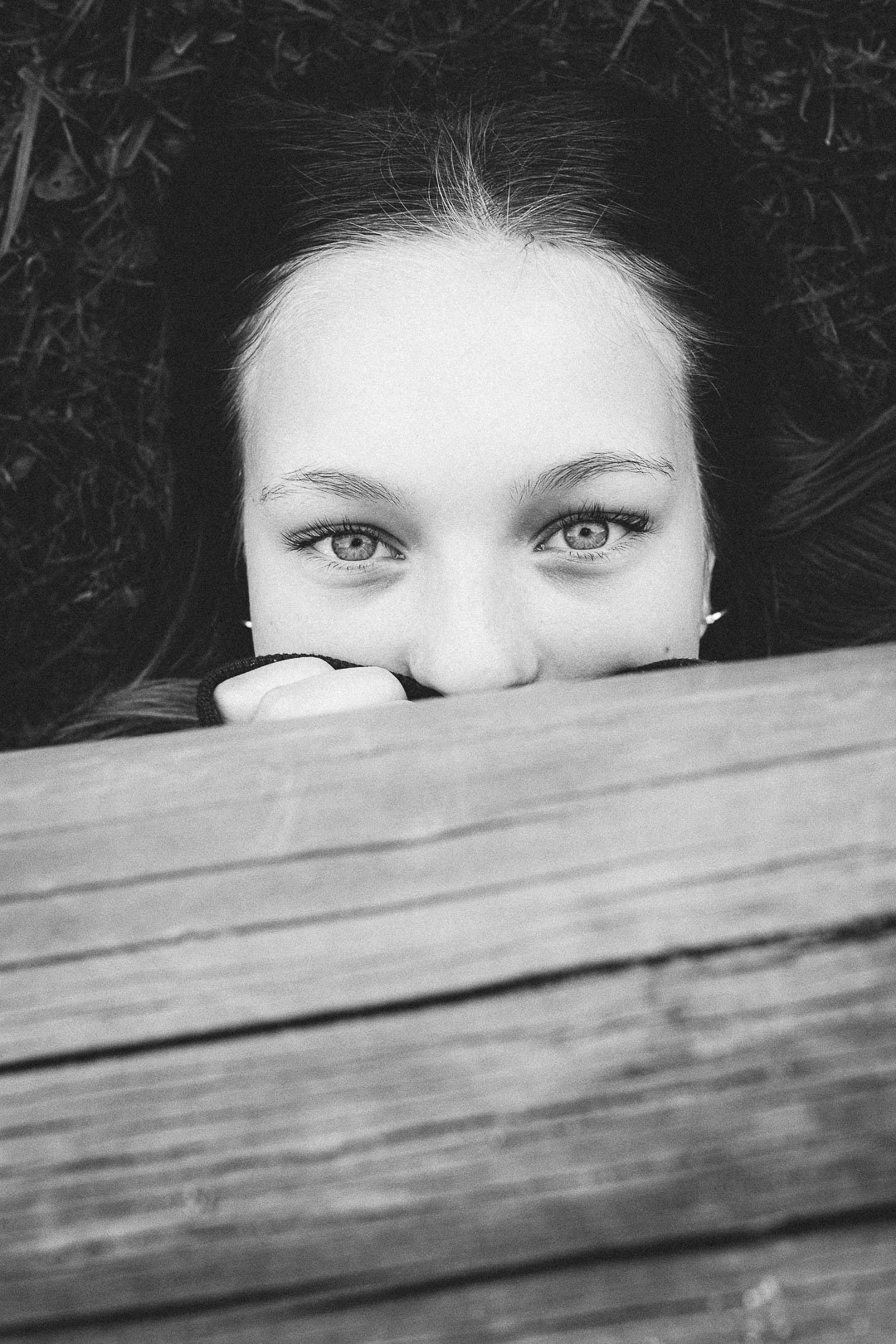 A black and white photo of a woman lying on her back with her face partially hidden behind a wooden surface, revealing only her eyes and forehead, with her hair spread out around her.