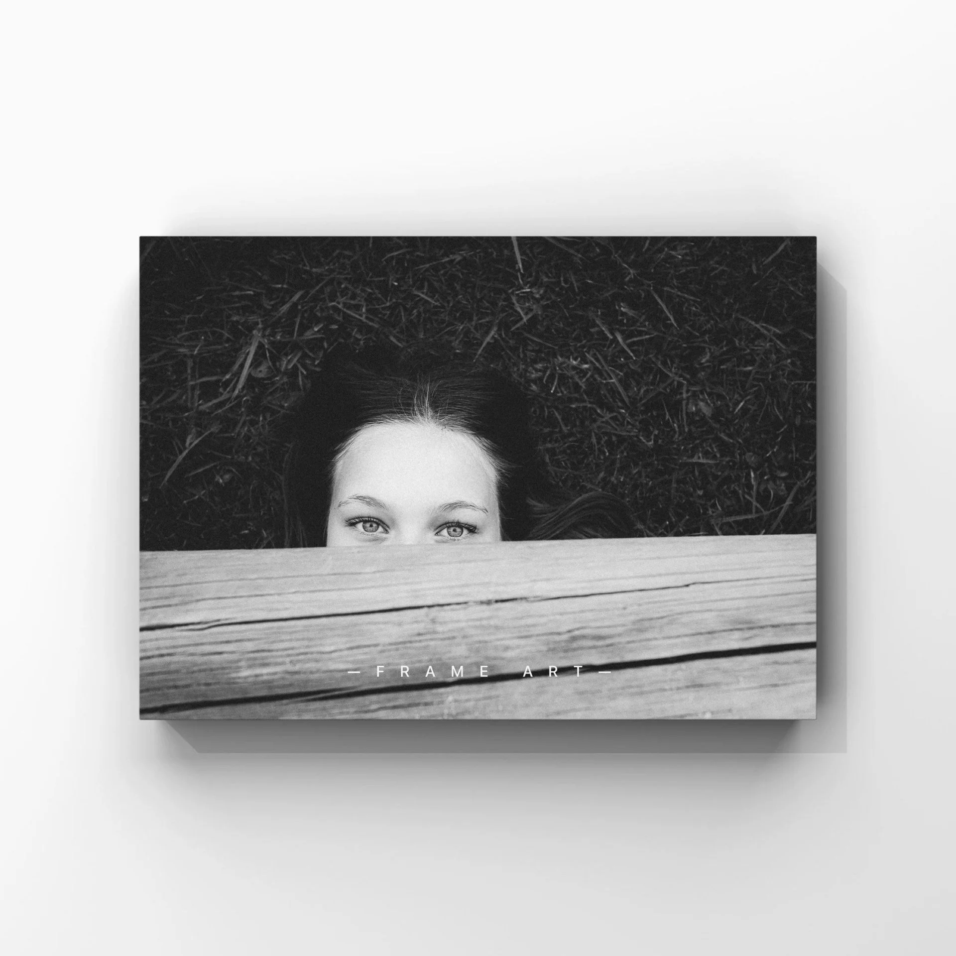 Black and white photograph of a woman with wide eyes peeking over a wooden plank, lying on the ground covered in grass or moss, with the words 'FRAME ART' below.