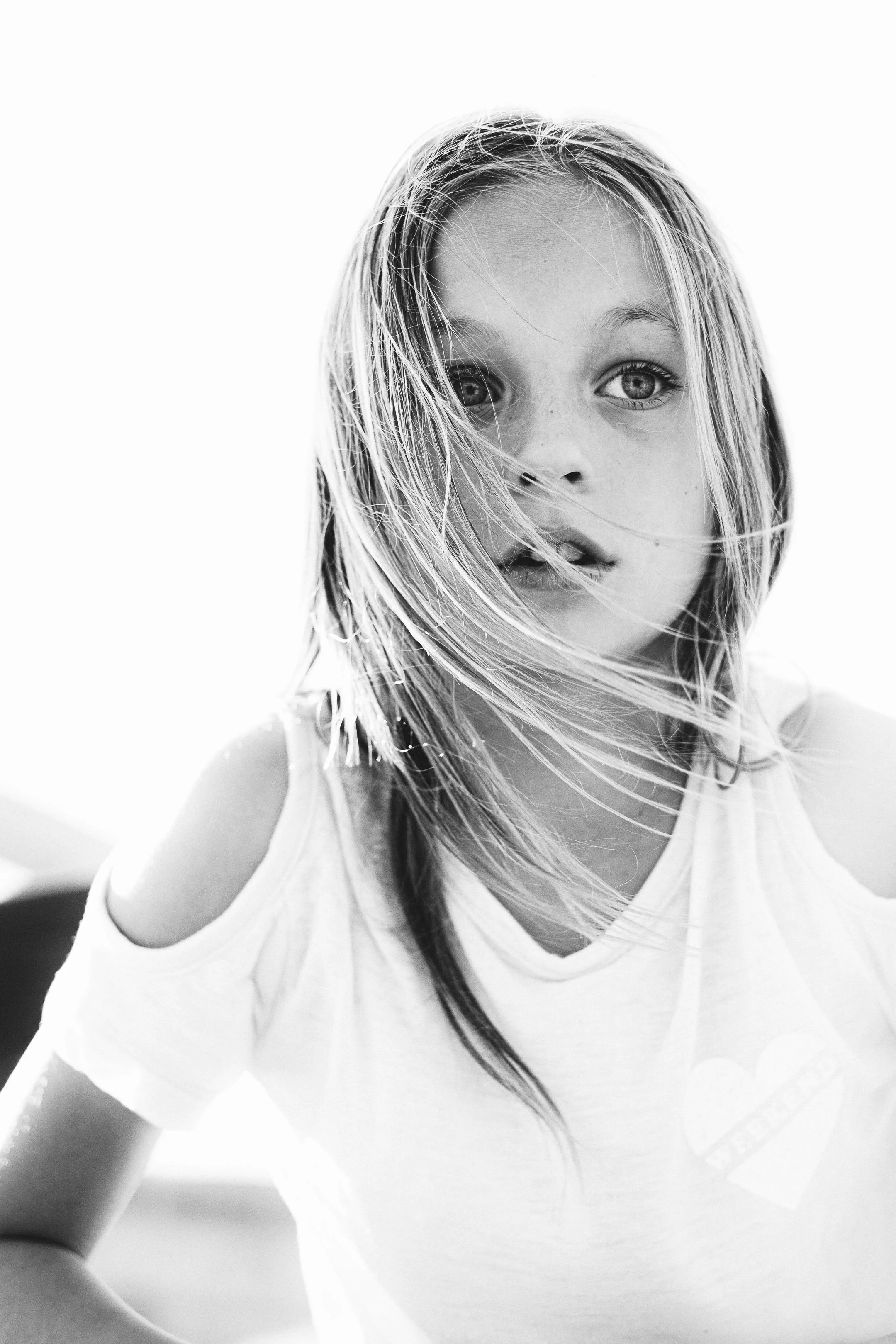 Black and white photo of a young girl with messy hair, wearing a white t-shirt with cut-out shoulders and a heart-shaped graphic on the chest, looking directly at the camera.